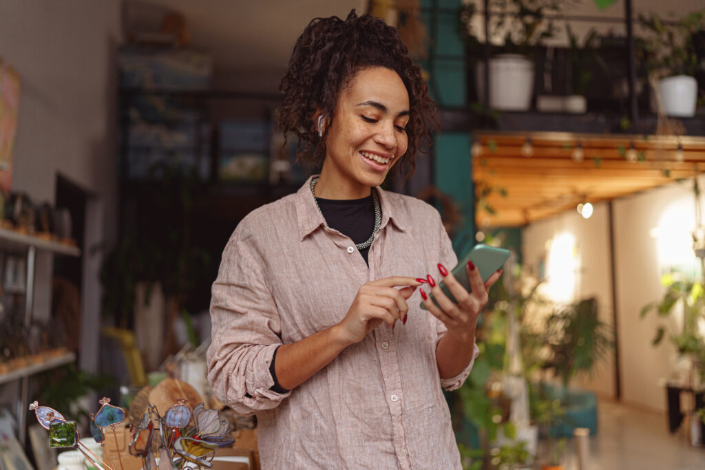 A woman smiling while using her smartphone in a cozy shop with plants, eyeglasses on display, and real estate chat open.