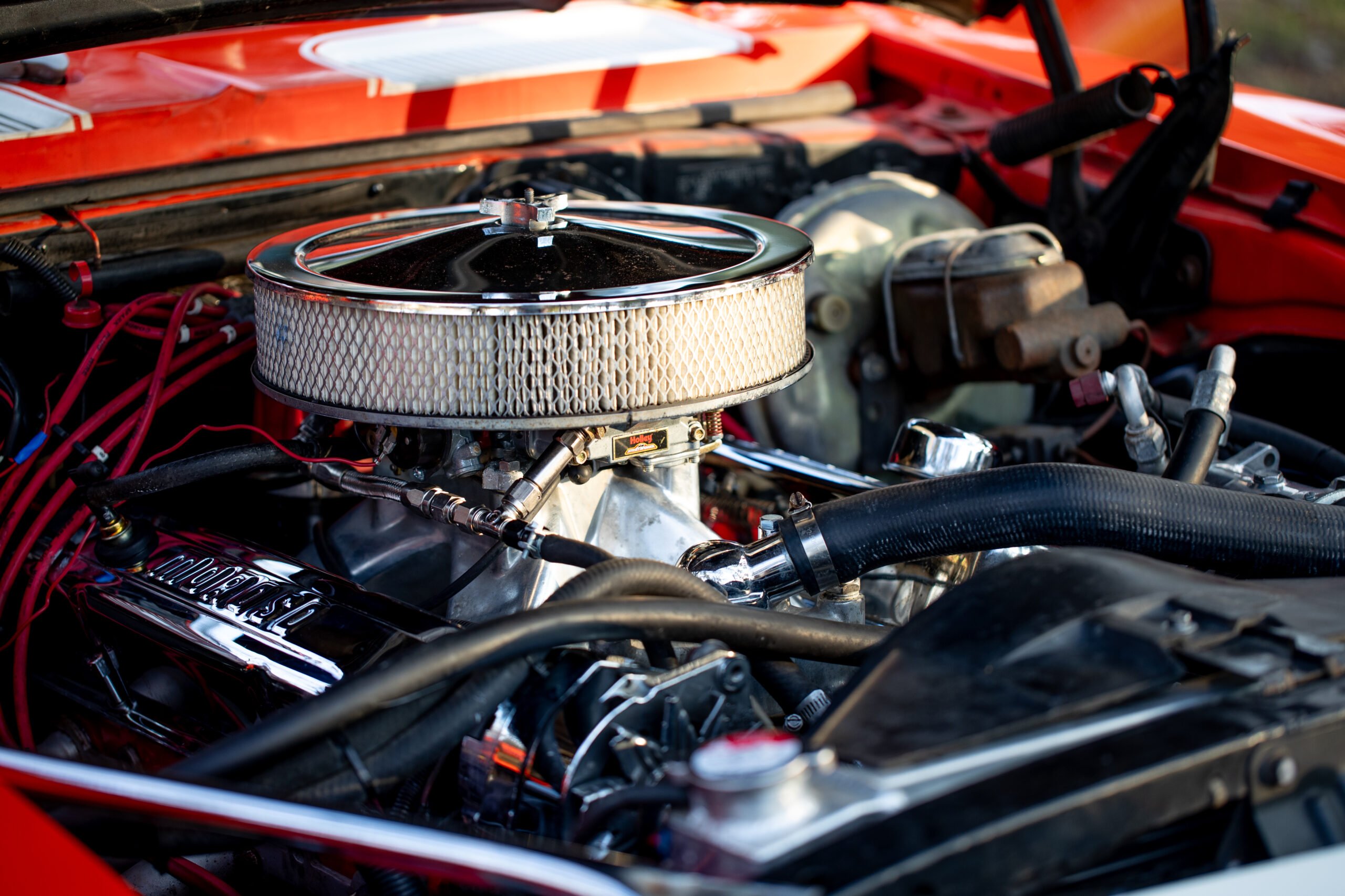 Close-up of a car engine with an air filter, visible parts, and wiring under the hood of a red vehicle.