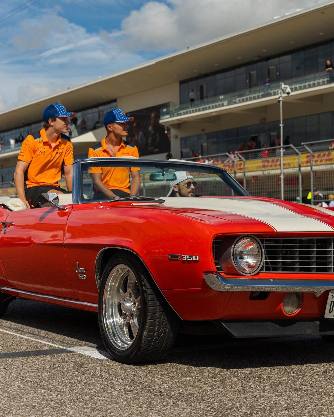Two people in orange shirts and blue caps standing next to a classic red Chevrolet Camaro SS at a racing event organized by Austin Realtor.