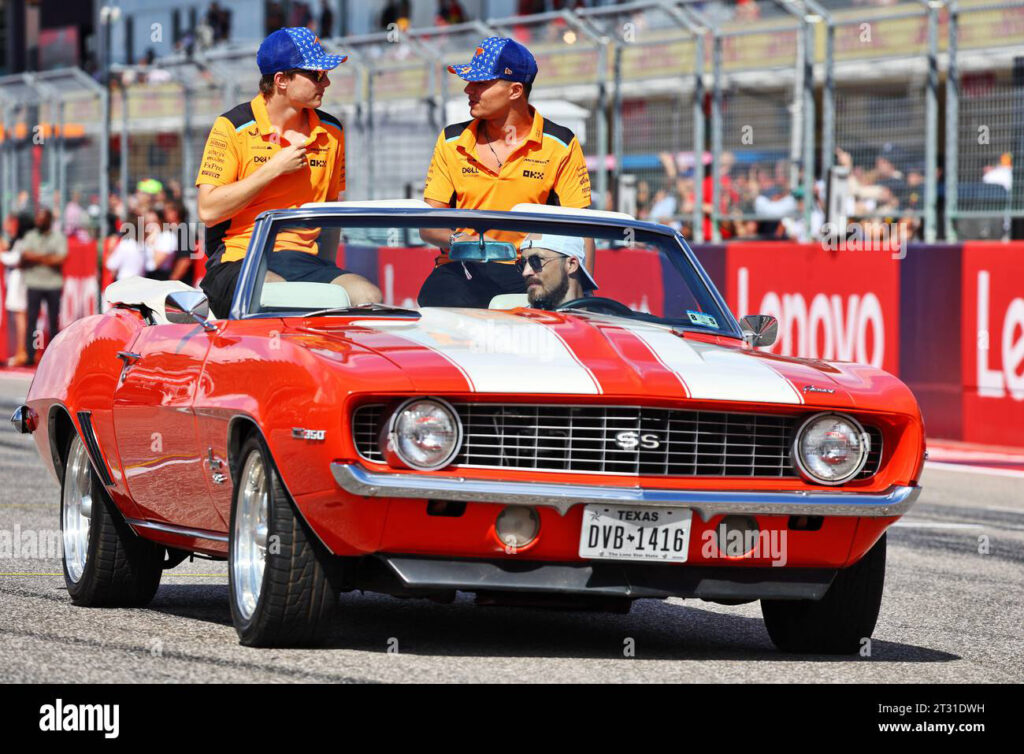 Two individuals in real estate racing team uniforms riding in a red vintage convertible car during a public home buying event.