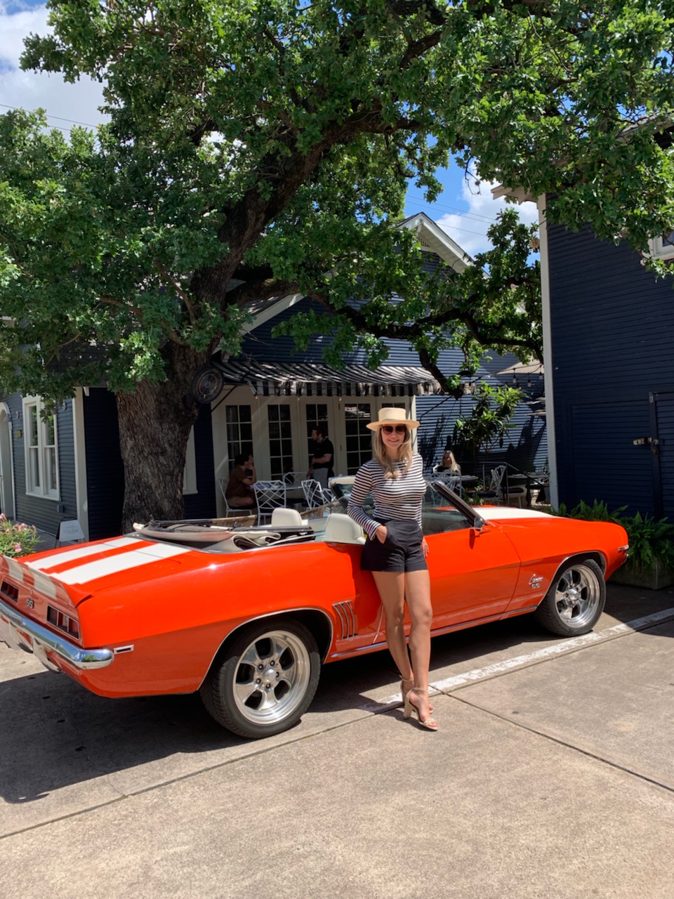 Woman standing beside an orange convertible car under a tree, showcasing her success as a top Austin Realtor.