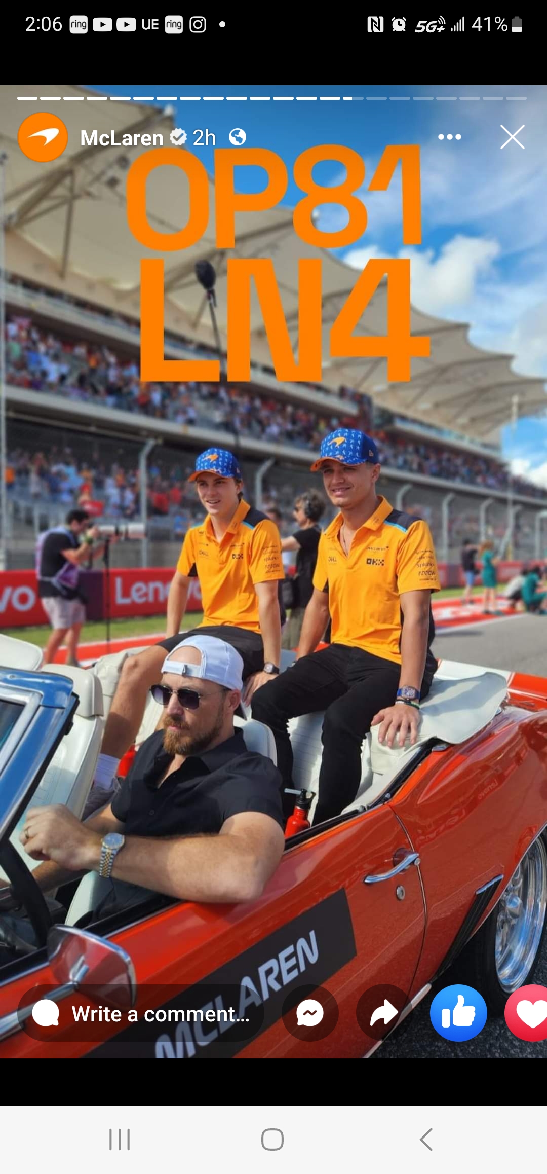 Two individuals in orange McLaren team apparel, engaging in a real estate chat, seated in a classic convertible car at a racing event.