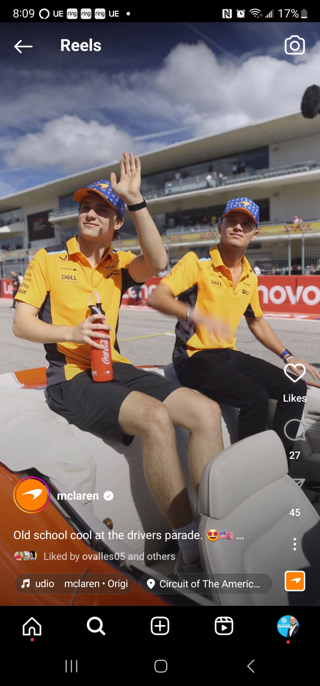 Two McLaren F1 team members, clad in team colors, participate in a drivers parade in Austin, waving at the crowd.