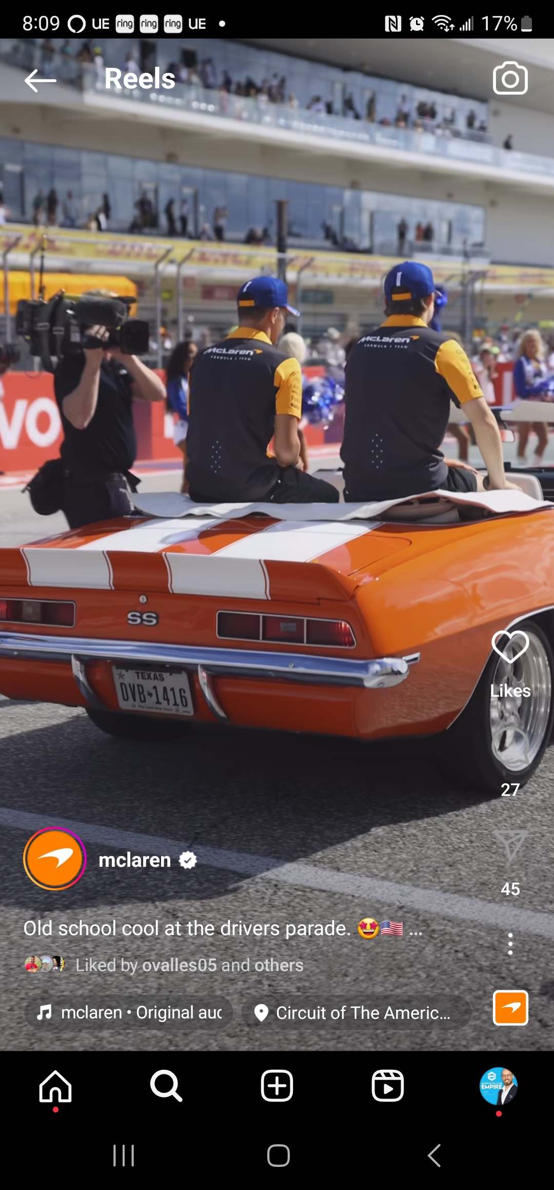 Two individuals in racing team apparel sitting on the back of a classic orange muscle car in a parade setting, discussing their next real estate chat.