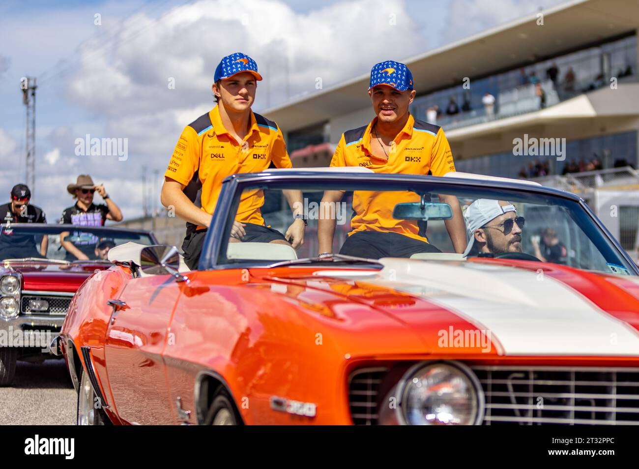 Two individuals in matching orange attire riding in a classic convertible car during a sunny real estate parade event.