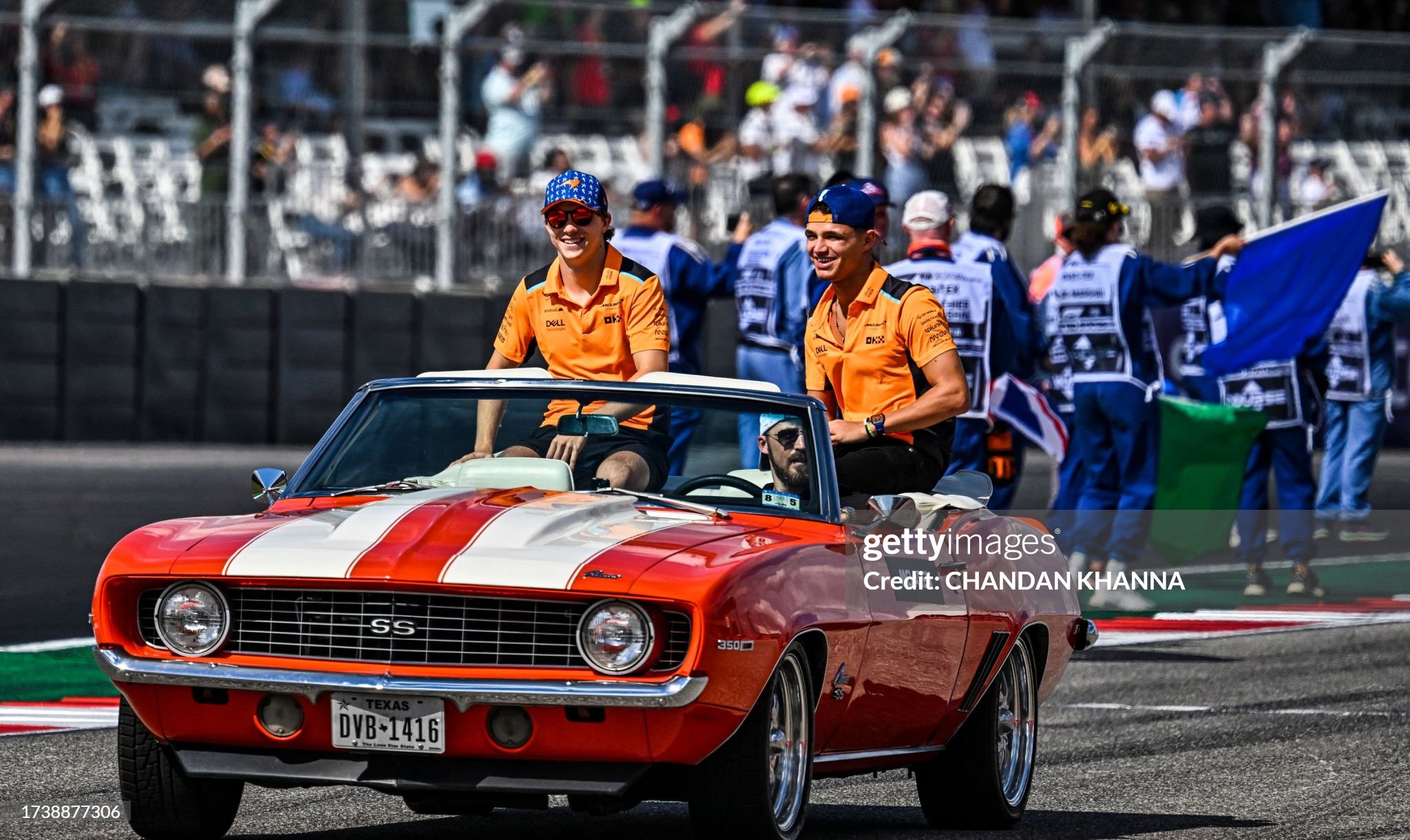 Two individuals in racing team attire riding in a vintage convertible car at a racetrack event organized by an Austin Realtor.