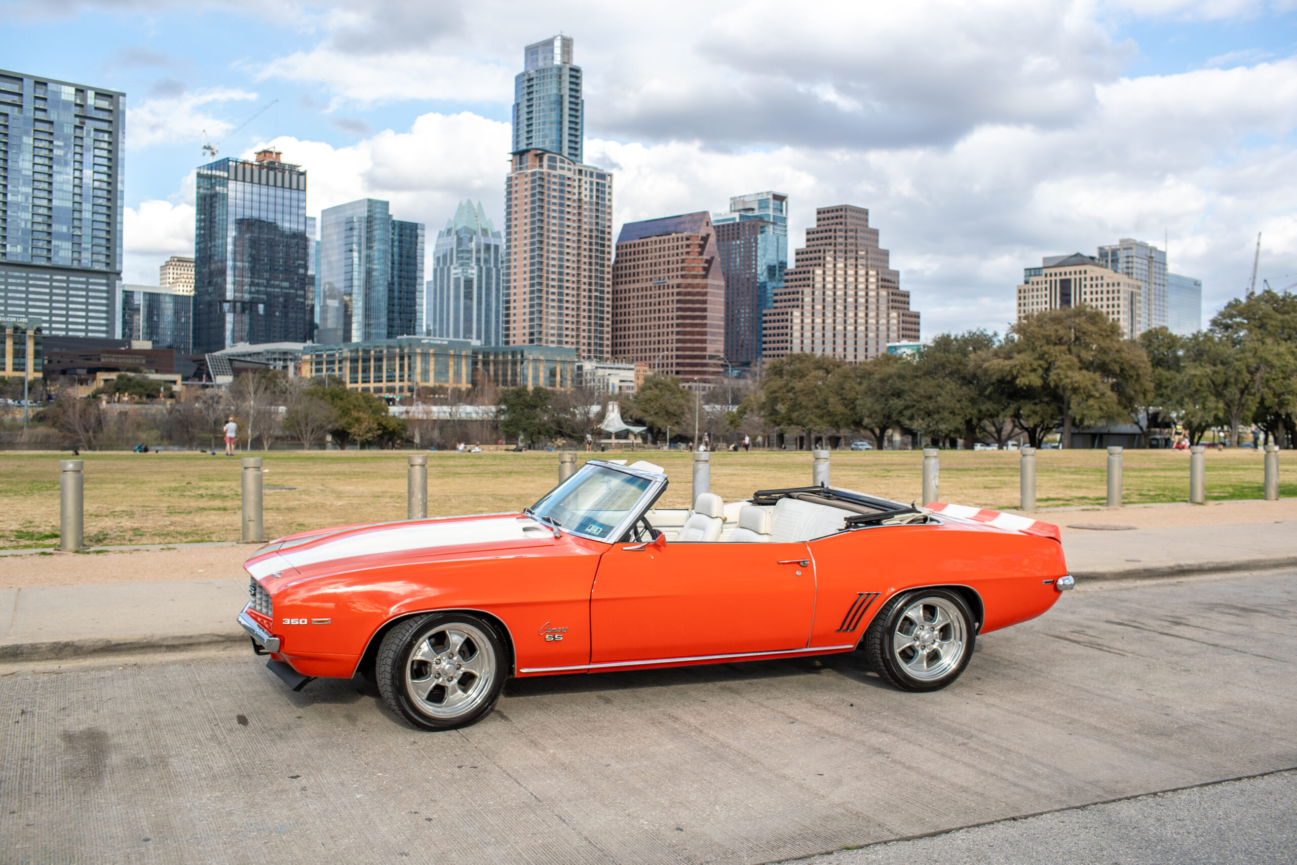 A bright orange convertible car parked near a grassy area with a city's skyline featuring several tall buildings in the background.