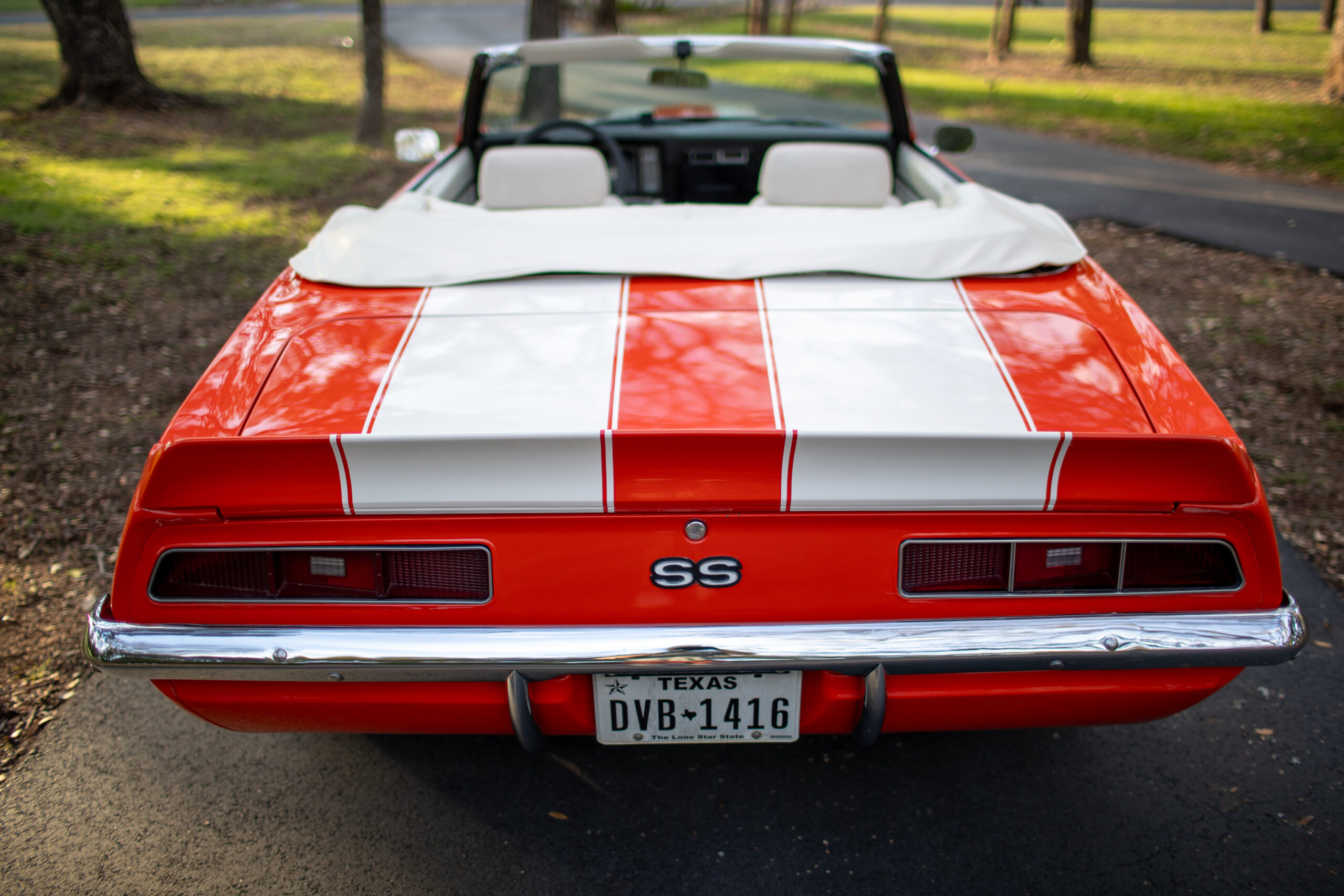 Rear view of a red and white convertible sports car with a Texas license plate "DVB-1416," parked on a driveway in a wooded area. The car has "SS" branding on the trunk.