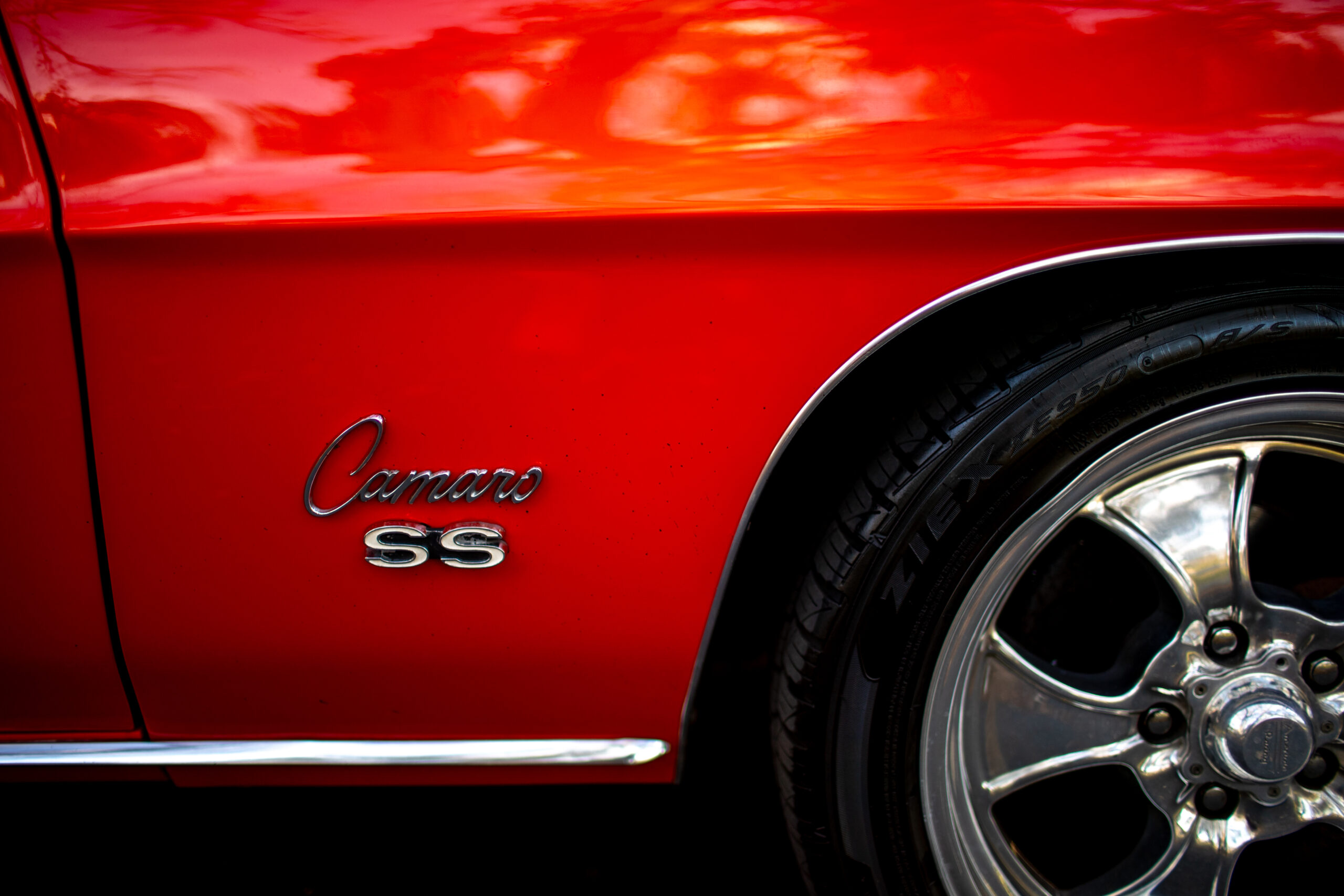 Close-up of a red Camaro SS showing the car's logo and part of a chrome-wheel with a shiny reflection.