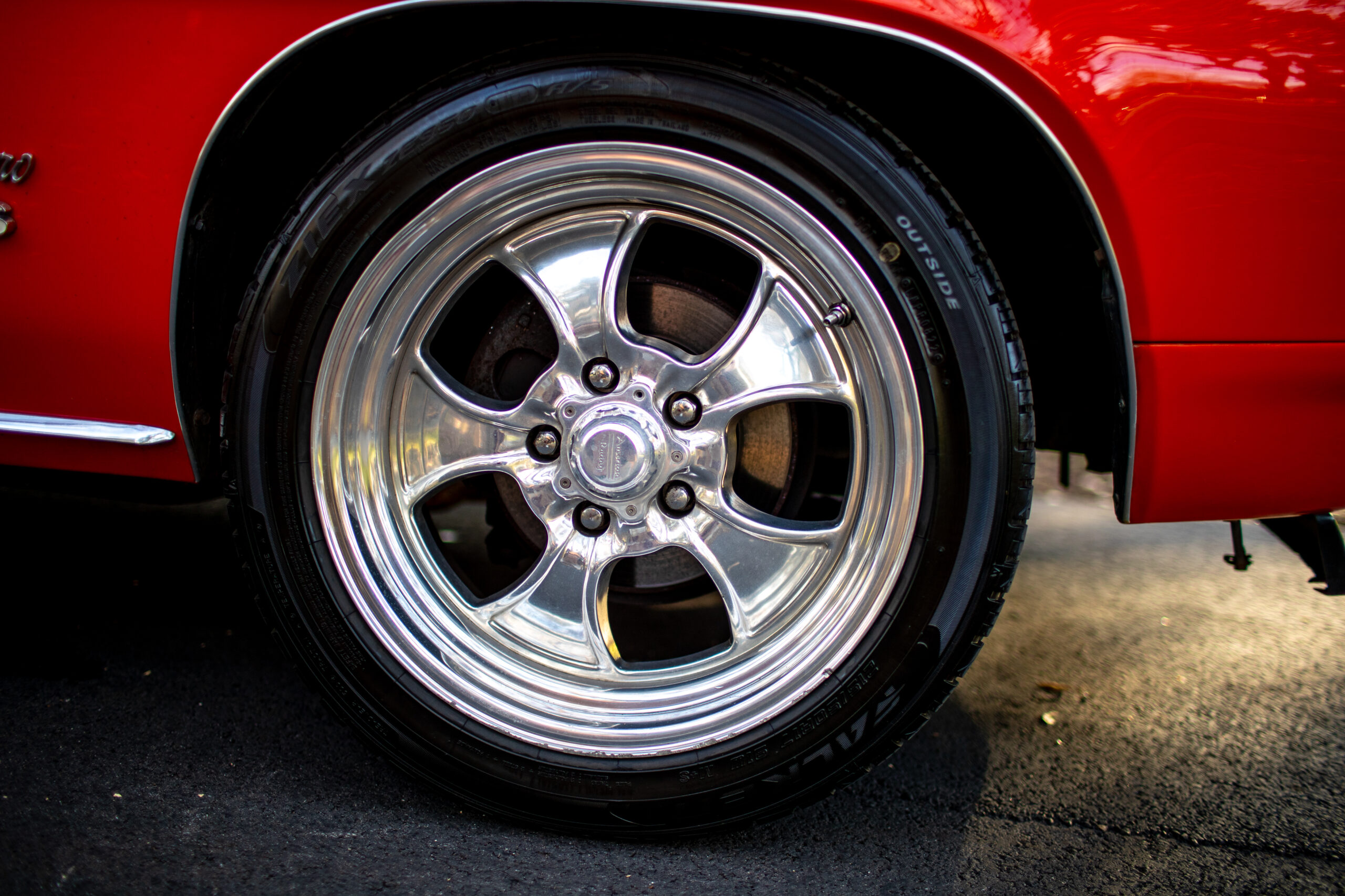 Close-up of a shiny chrome alloy wheel on a red car, mounted on a black tire. The car's exterior paint is visible in the background.