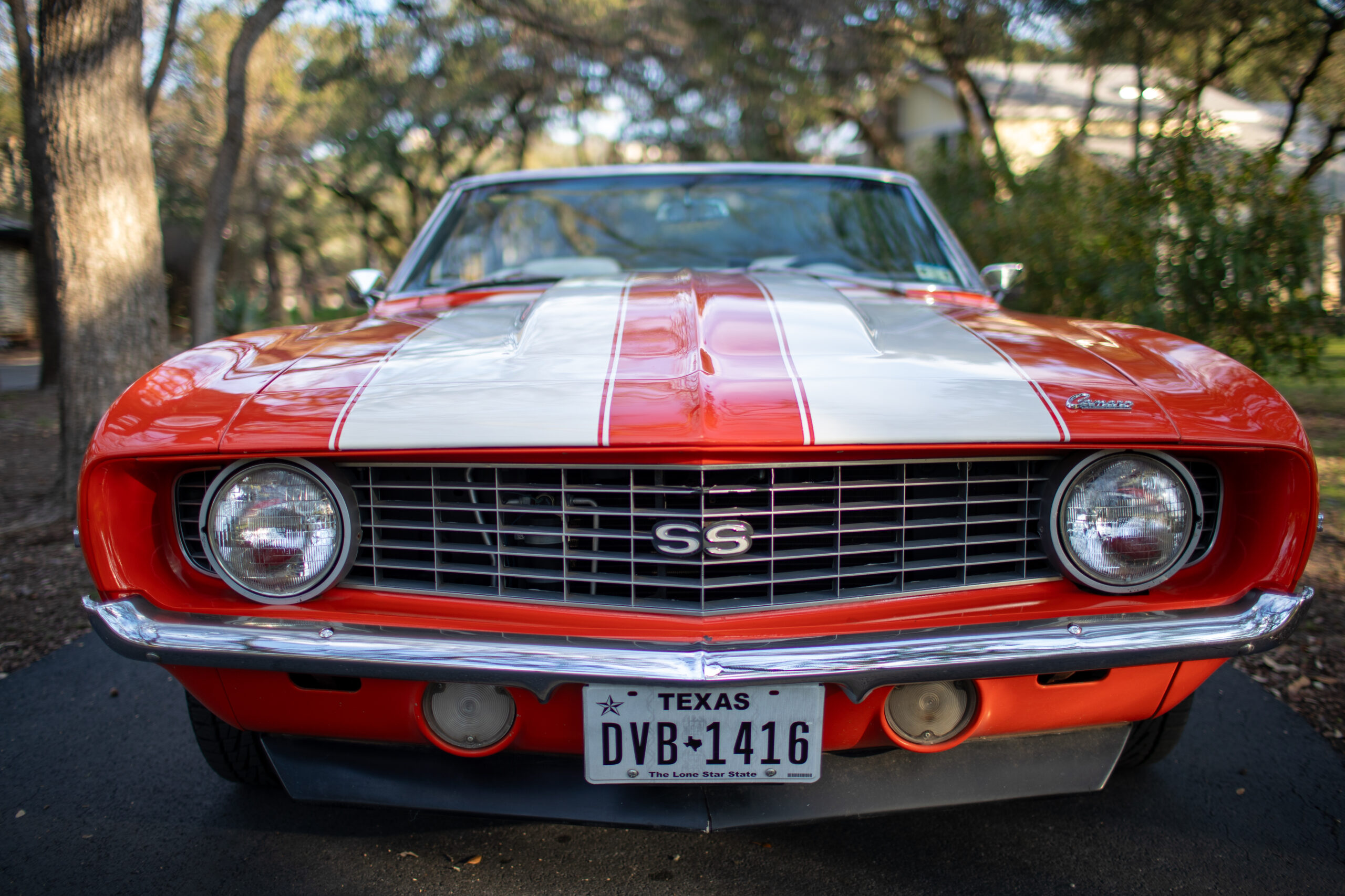 Front view of a vintage red Chevrolet Camaro with white racing stripes, parked on a driveway. Texas license plate "DVB-1416". Trees and sunlight in the background.