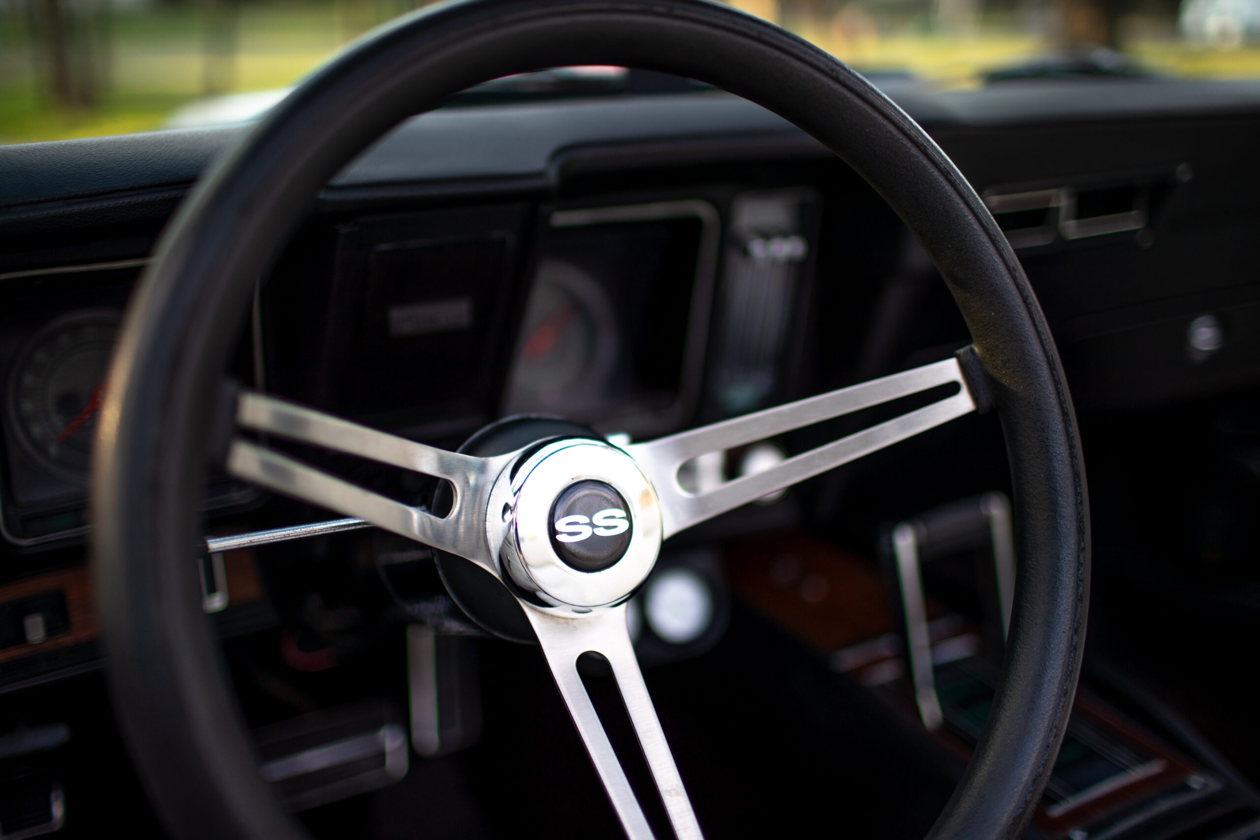 Close-up of a classic car's steering wheel with an "SS" emblem at the center, featuring a black and chrome design. The car's dashboard and gauges are visible in the background.