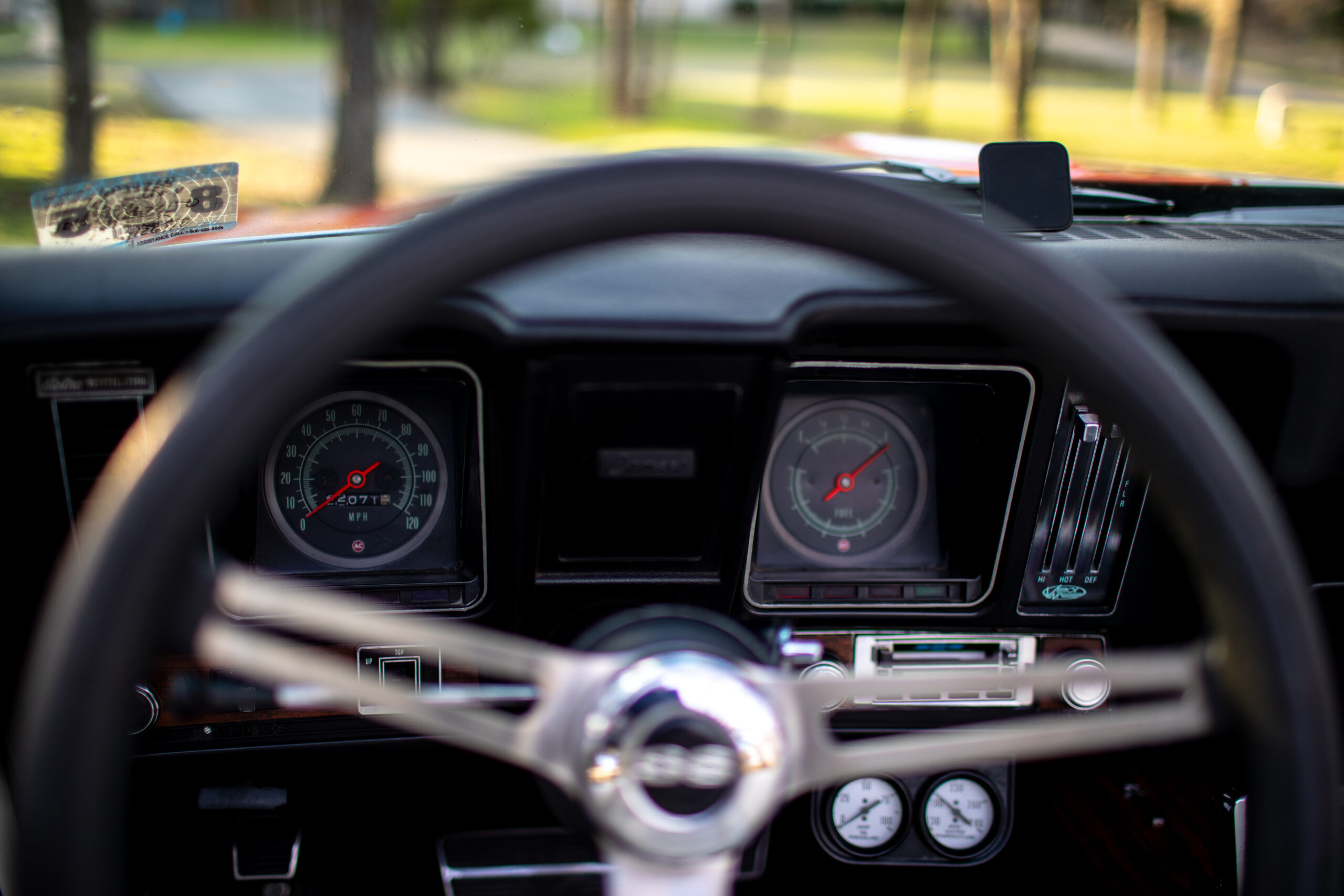 Interior view of a car dashboard displaying speedometer, tachometer, and other gauges behind the steering wheel, with a blurred background observed through the windshield.