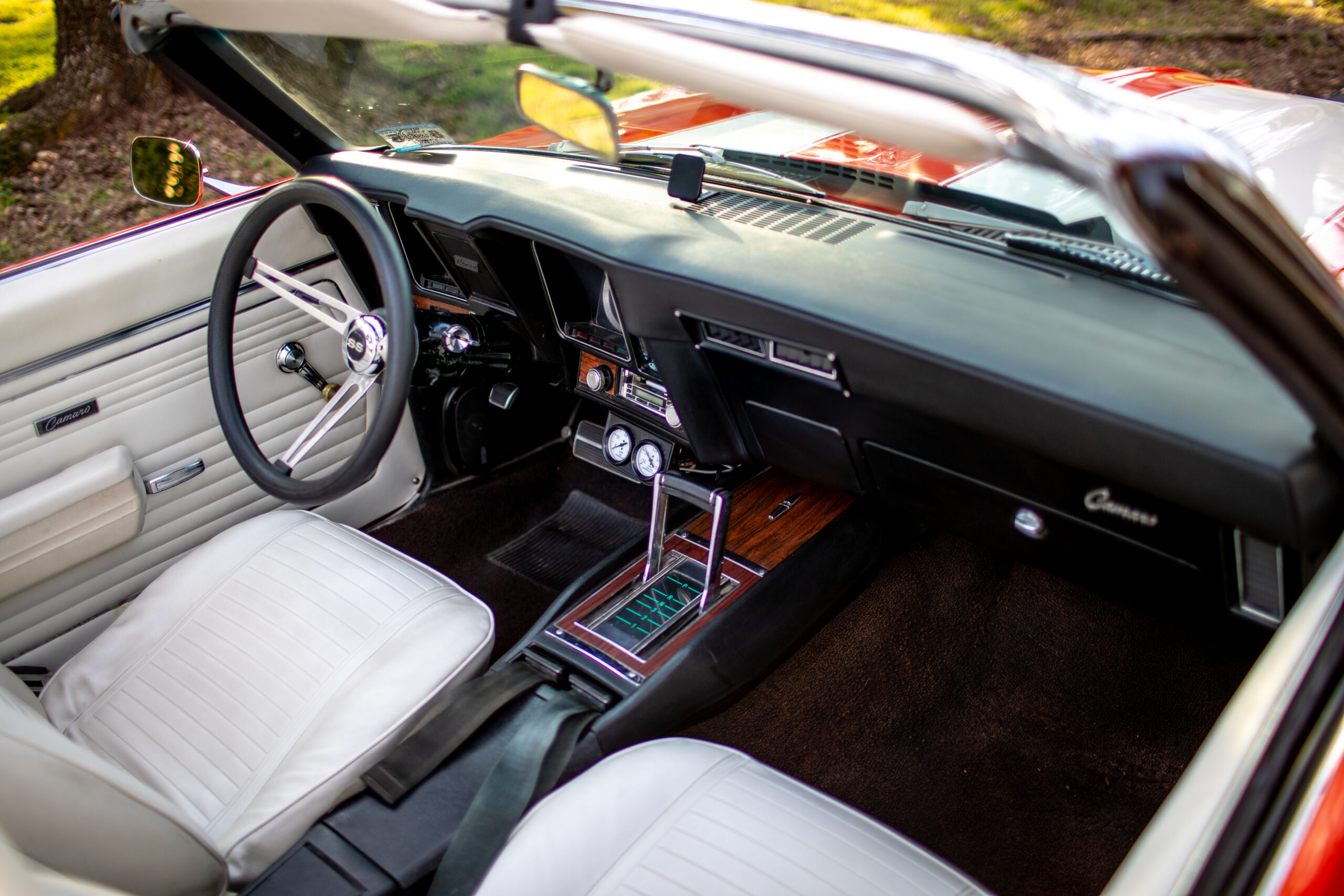 Interior view of a classic convertible car with white leather seats, a black dashboard, and wood accents. The view shows the steering wheel, gear shift, and control panel.