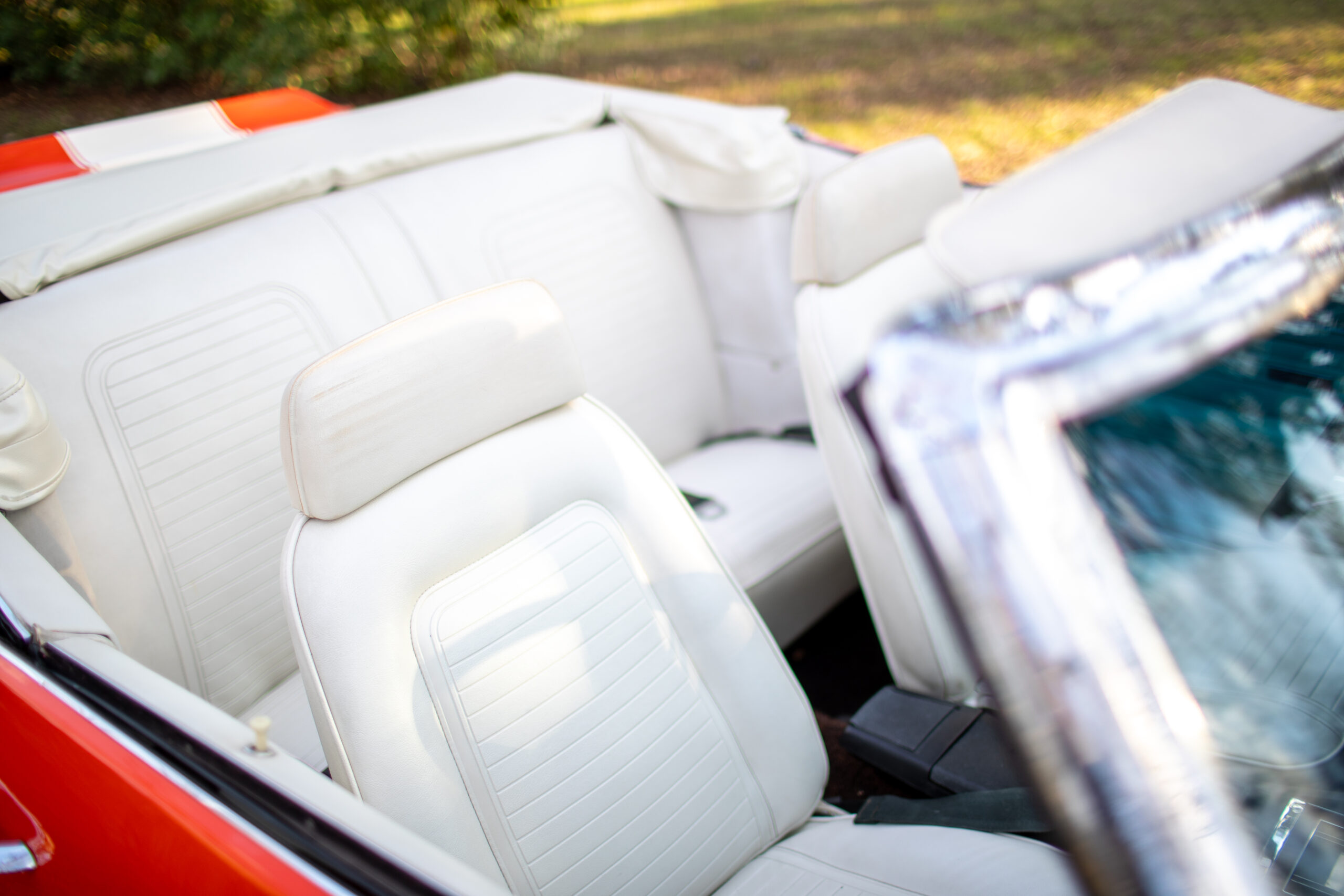 Close-up of the white leather interior of a red convertible car with the top down. The front seats are visible, with one headrest slightly tilted forward. Trees and grass are in the background.
