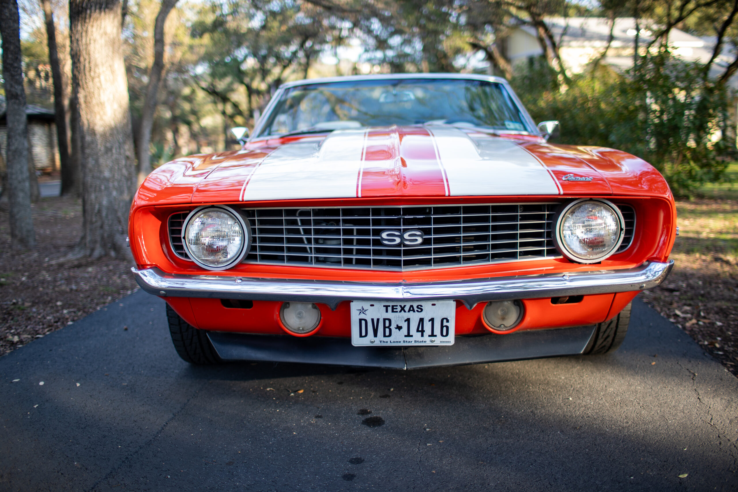 Front view of a red vintage muscle car with white racing stripes and a Texas license plate parked on a driveway.