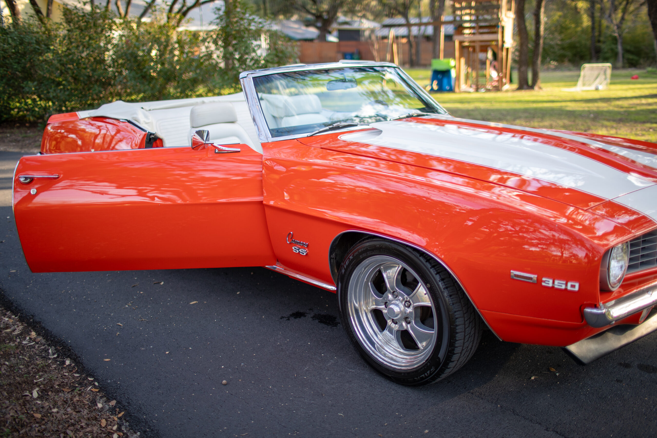 A bright orange vintage convertible car with white racing stripes has its driver's side door open. The car is parked on a paved surface with trees and a playset visible in the background.