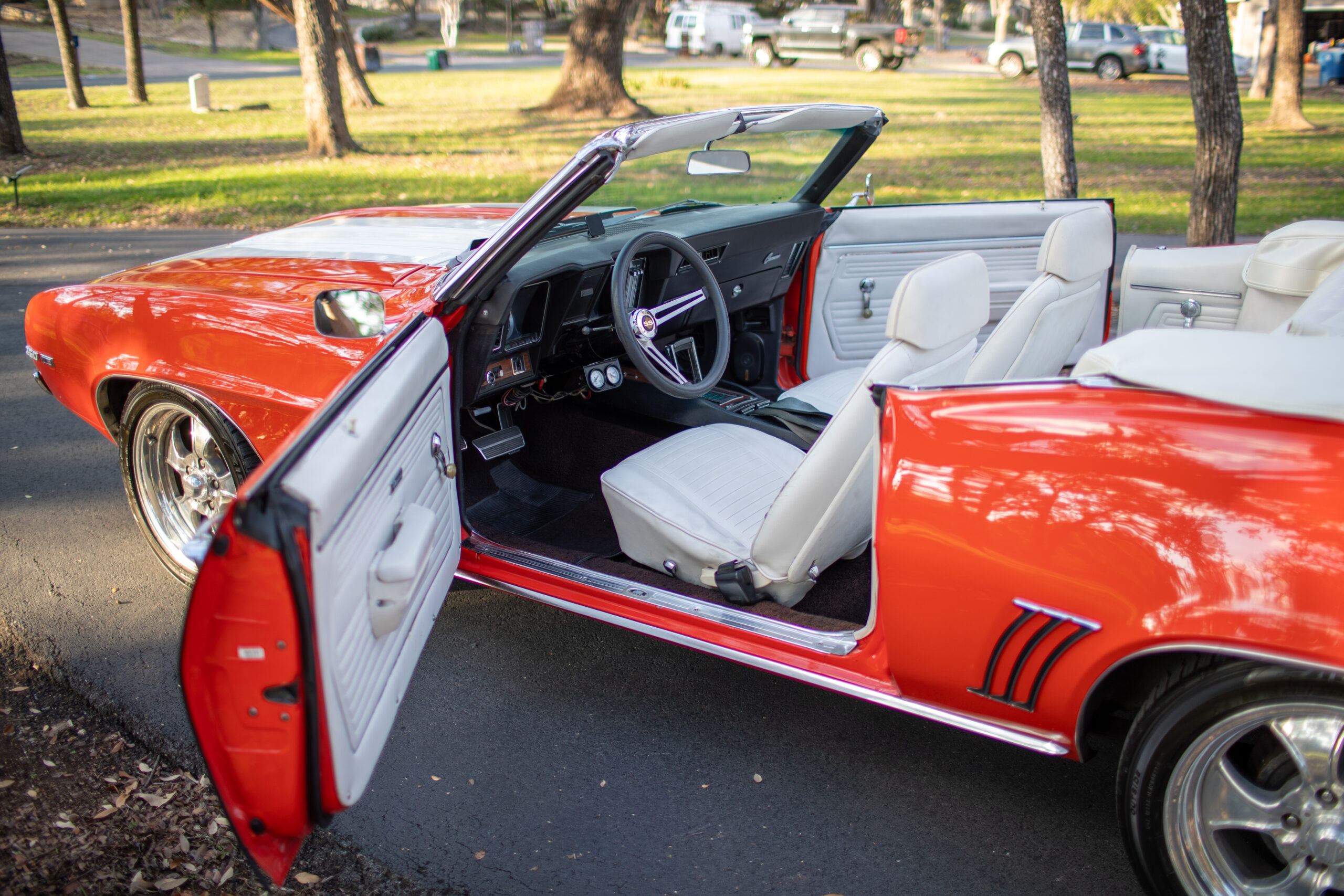 A red convertible car with its driver's side door open, revealing a white interior and a vintage dashboard, is parked on a paved road in a park with trees and other parked vehicles in the background.