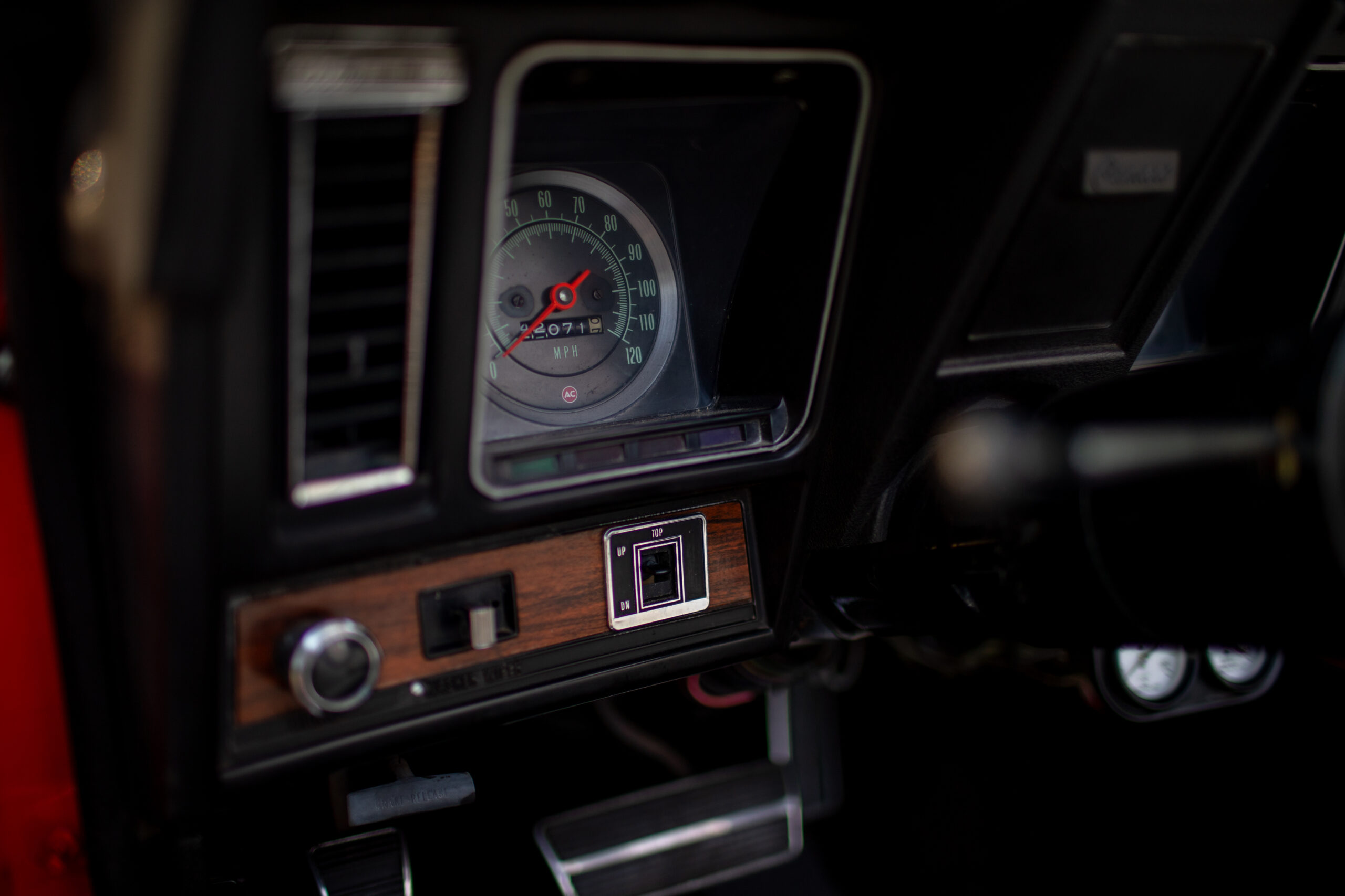 Close-up view of a classic car dashboard showing a speedometer with a red needle, a wooden panel with buttons and knobs, and parts of the steering wheel and pedal visible.