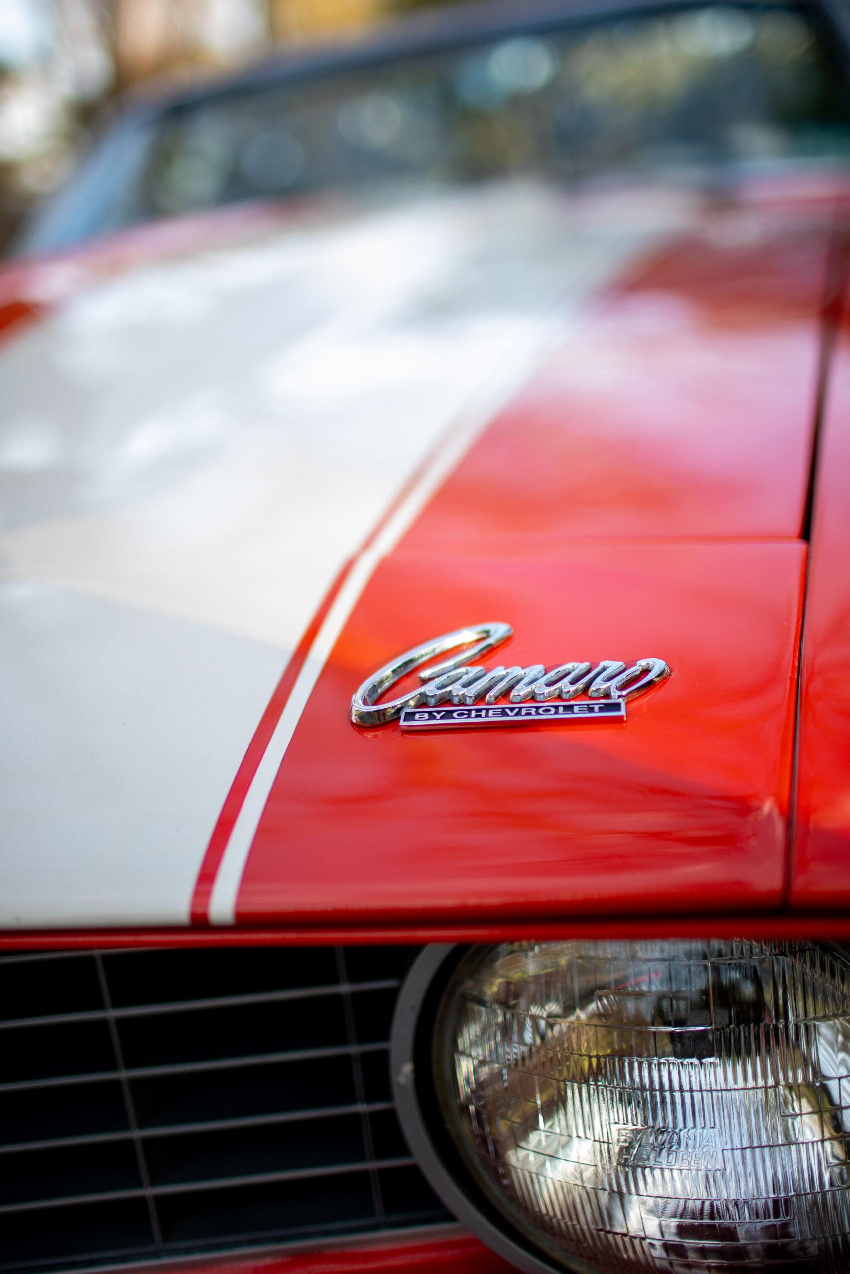 Close-up of a red and white classic Camaro car with a focus on the front emblem and headlight.