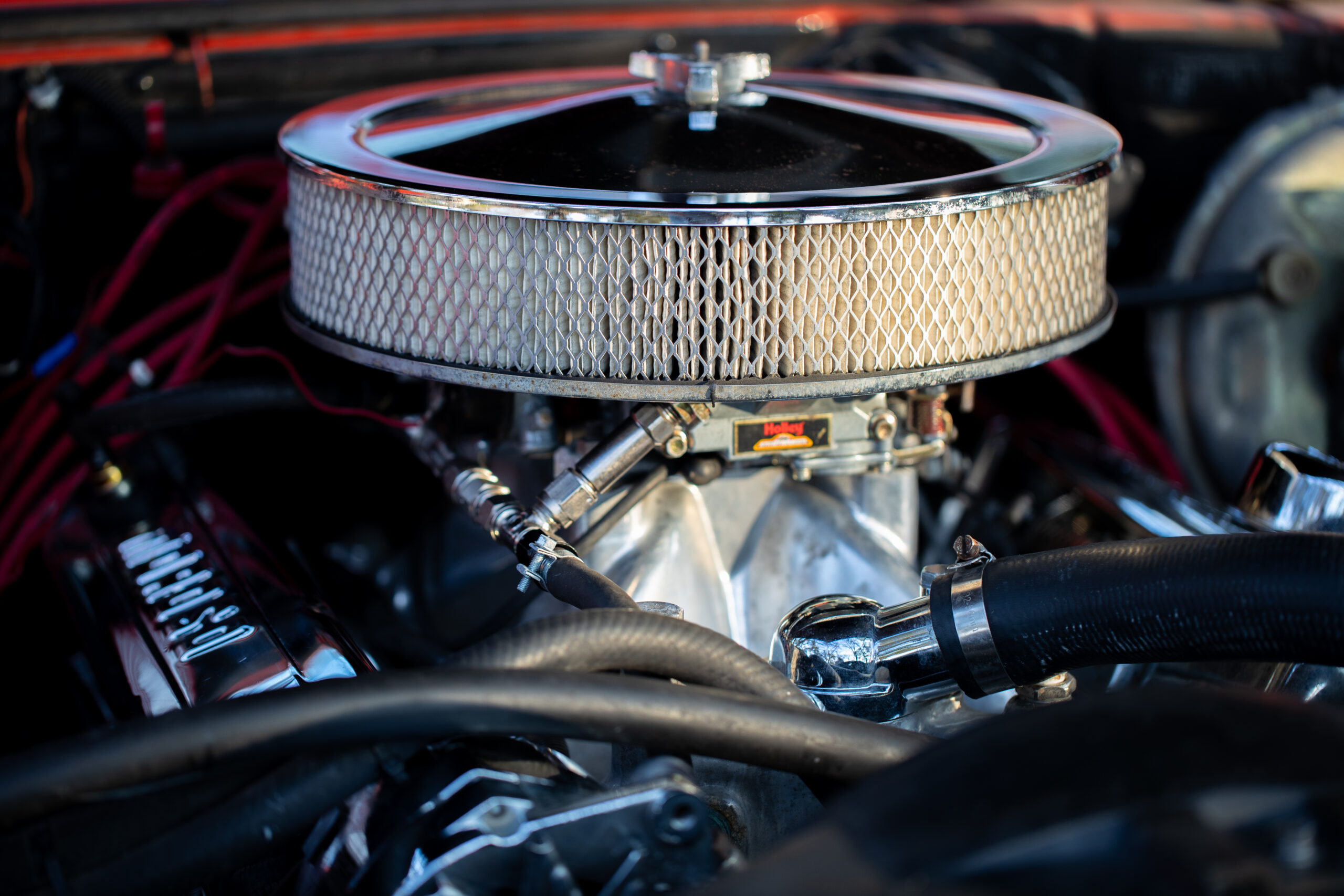 Close-up view of a car engine showing a round, chrome air filter with a mesh design mounted on top of the engine components. Various hoses and wires are visible around the engine.