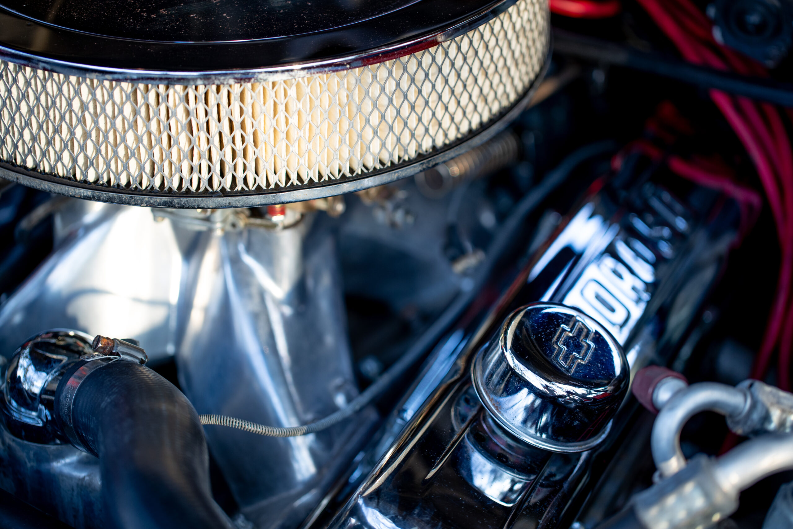 Close-up of a car engine, highlighting the air filter and chrome valve cover. The Chevrolet emblem is visible on the valve cover.