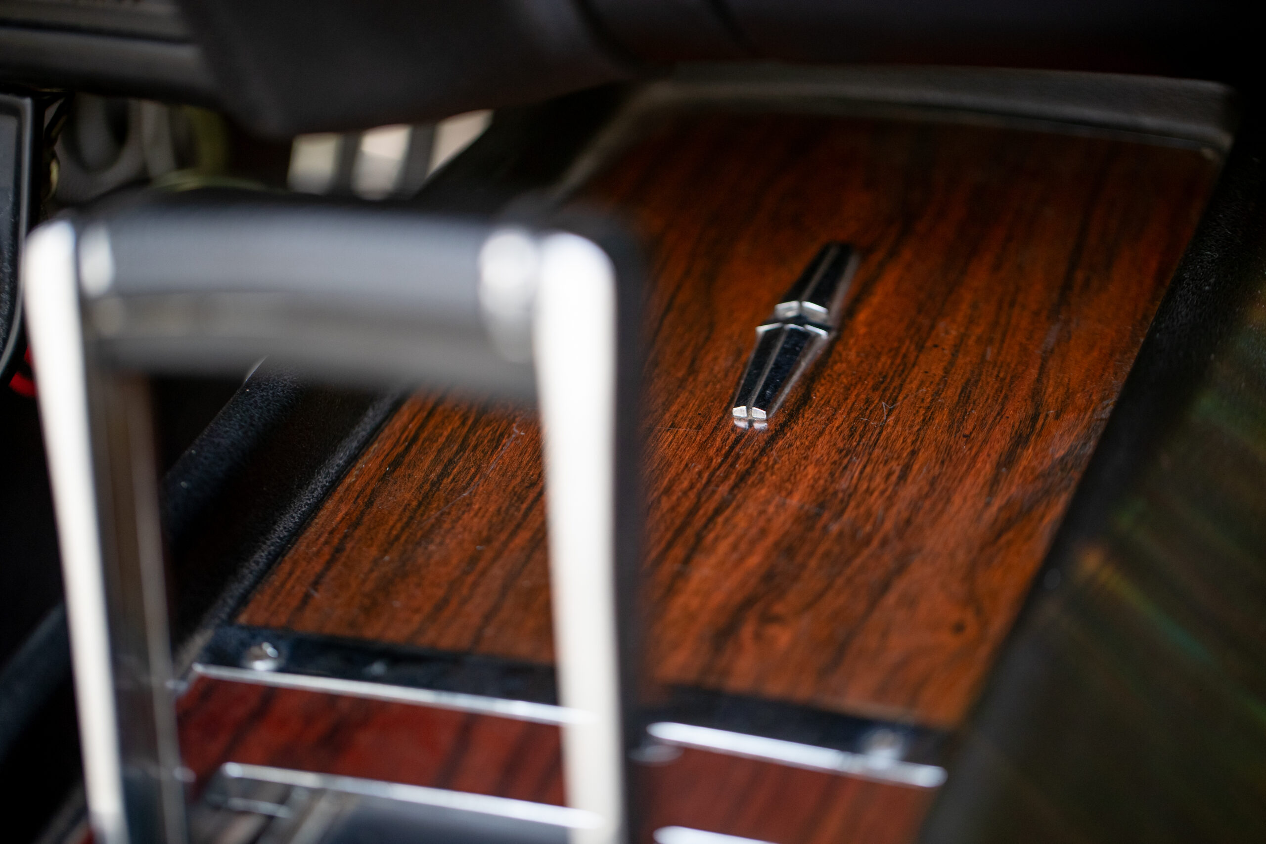 Close-up of a car's gear shift area featuring a wooden panel with a metallic emblem in the center, with part of the gear stick visible in the foreground.