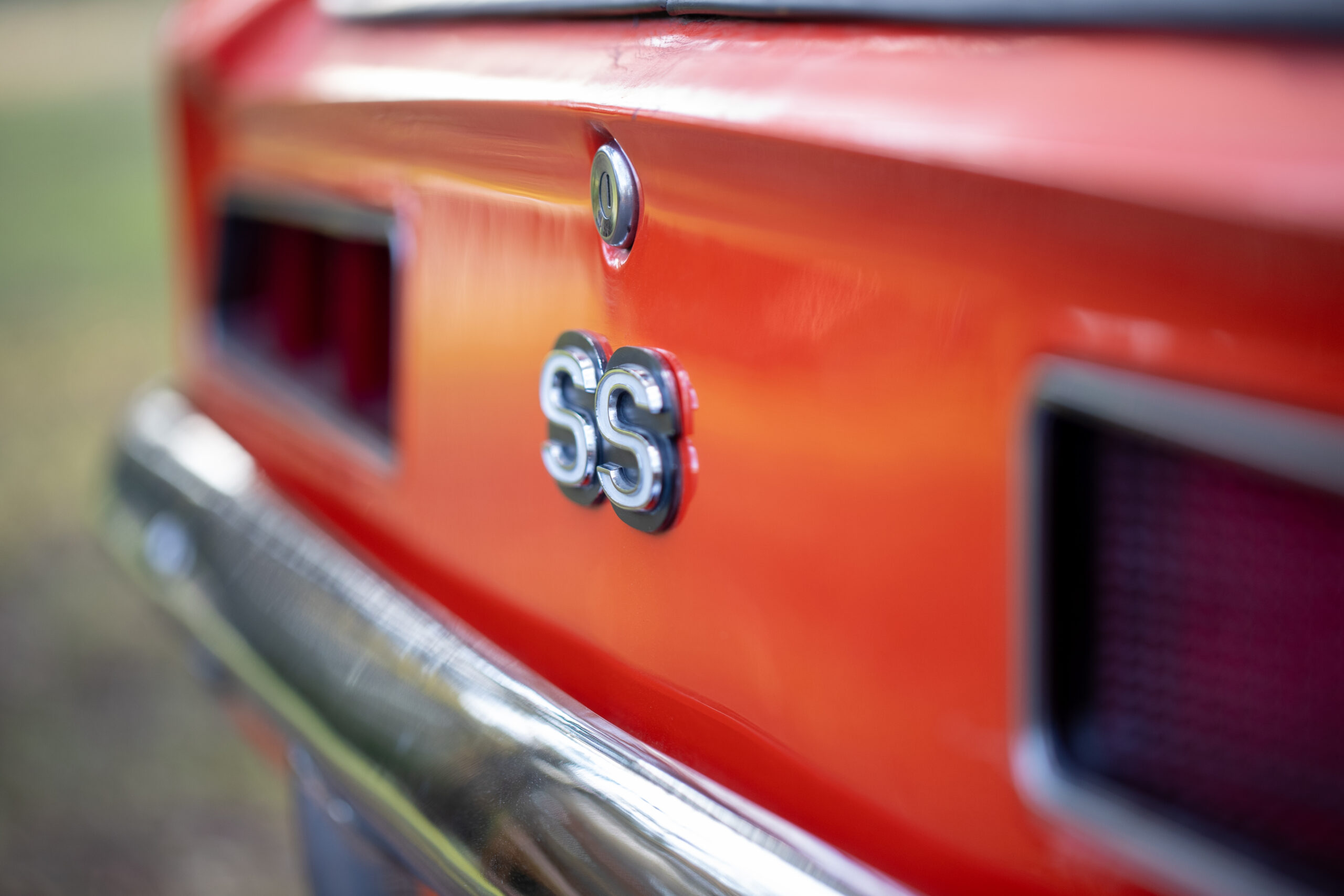Close-up of the rear of a vintage orange car with a chrome "SS" emblem visible, along with part of the rear bumper and taillight area.