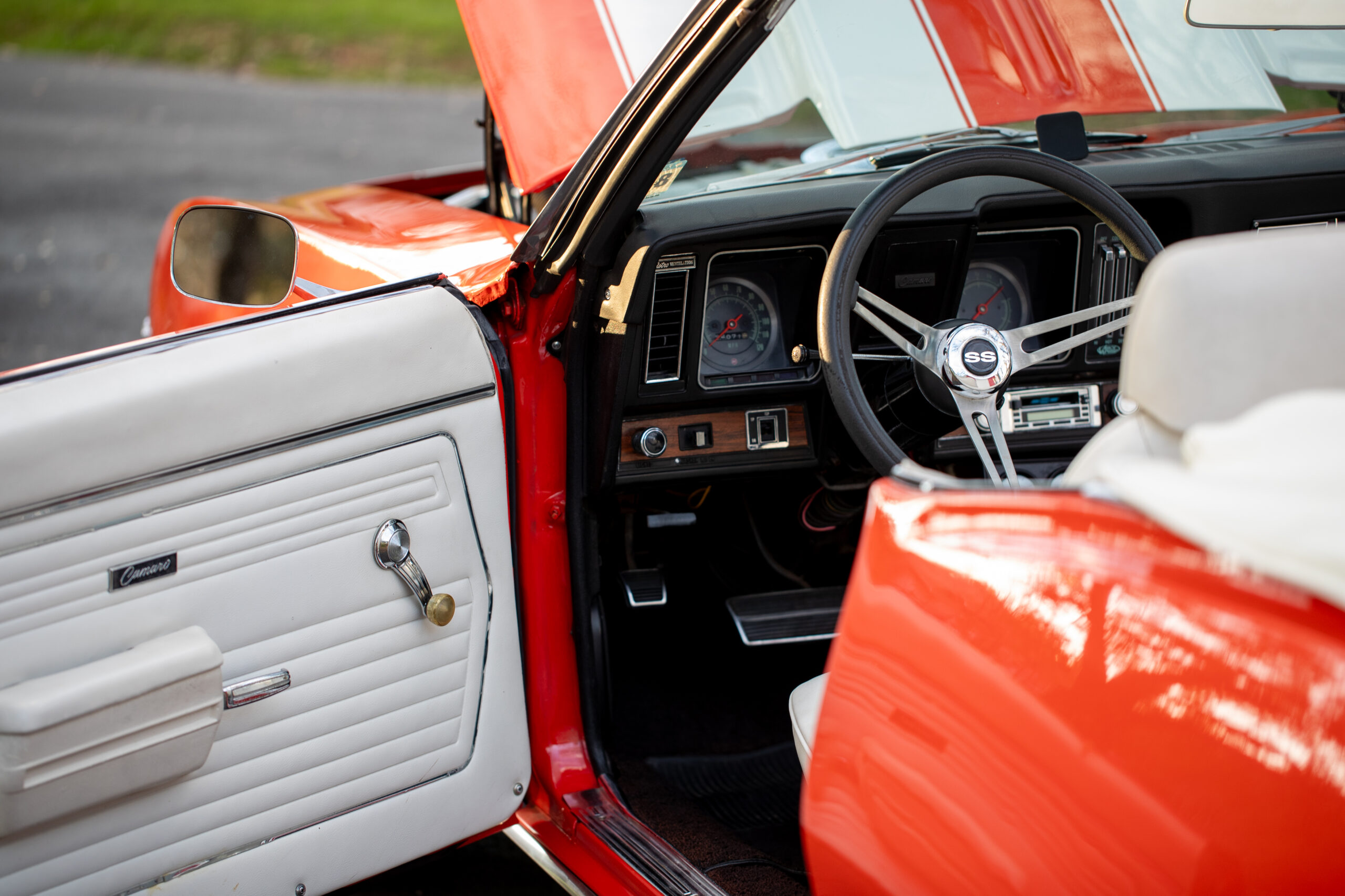 Interior of an orange classic convertible car with the driver's door open, showing white leather door panels, steering wheel, and dashboard with gauges.