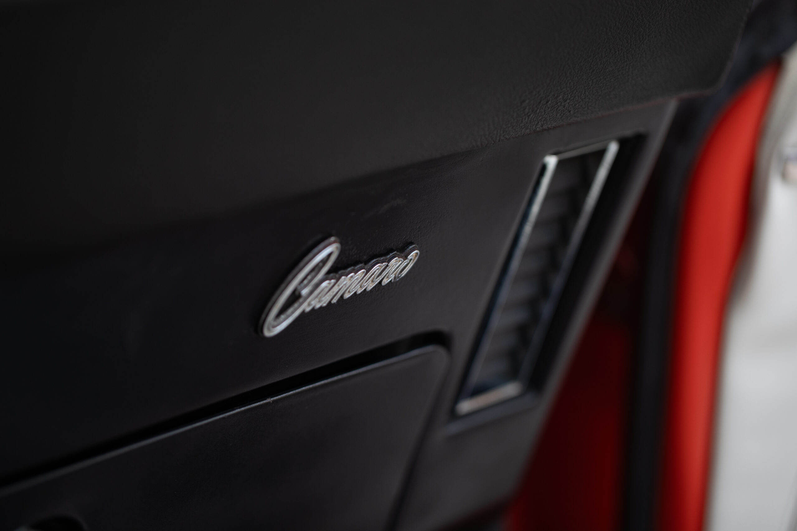 Close-up of a car interior showing the "Camaro" logo and a portion of the air vent. The dashboard appears to be black.