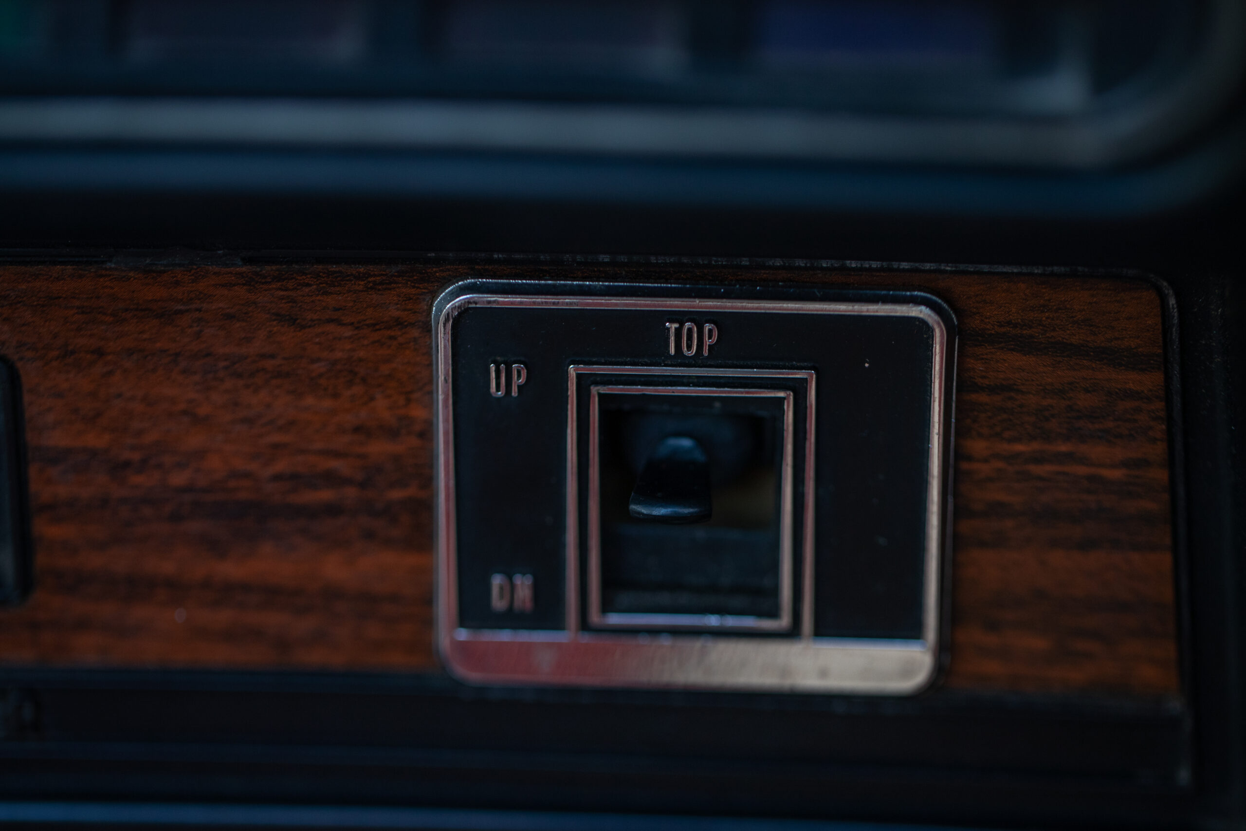 Close-up view of a car dashboard featuring a toggle switch for the convertible top, labeled "UP" and "DN," set against a woodgrain panel.