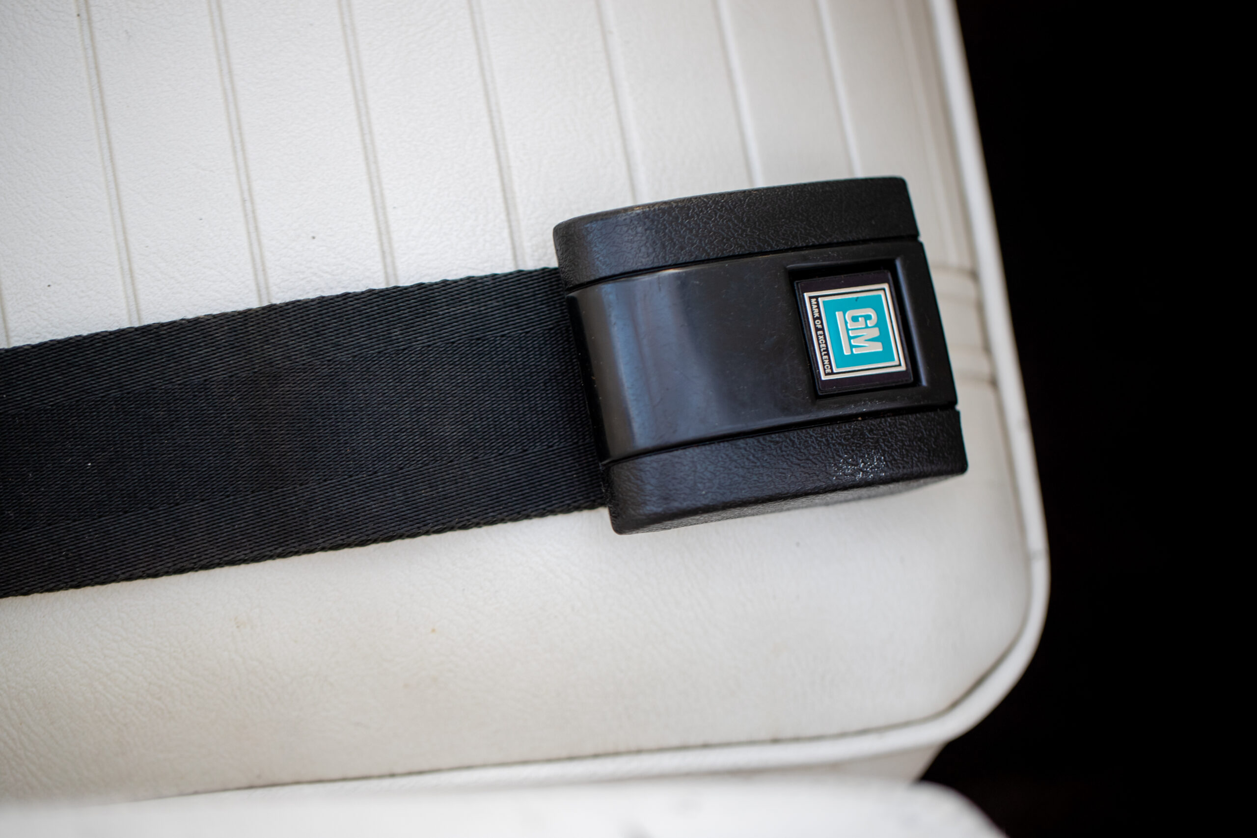 Close-up of a black seatbelt buckle with a GM logo, fastened on a white padded car seat.