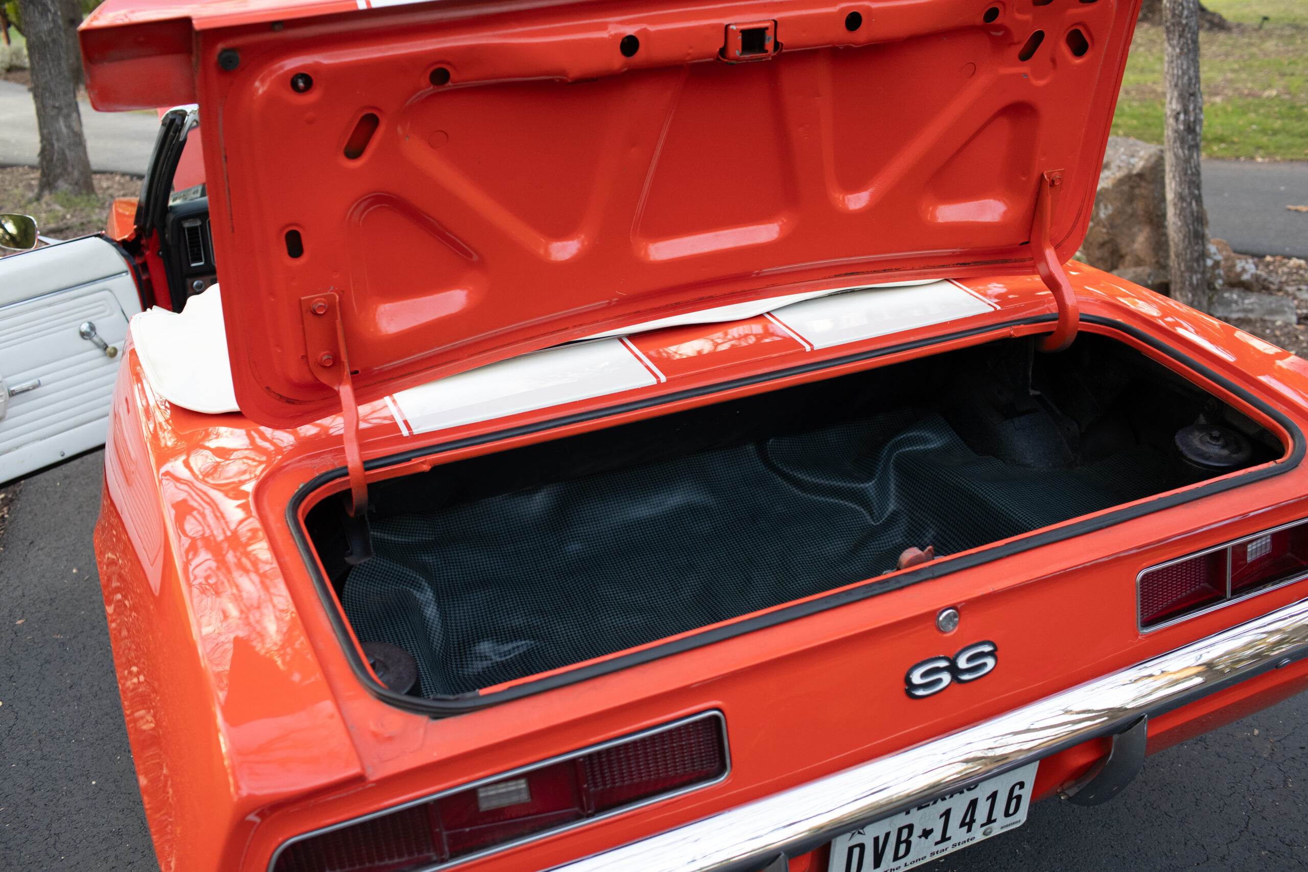 Open trunk of an orange vintage car with an SS badge, displaying a clean, empty cargo area. A white door and trees are visible in the background.