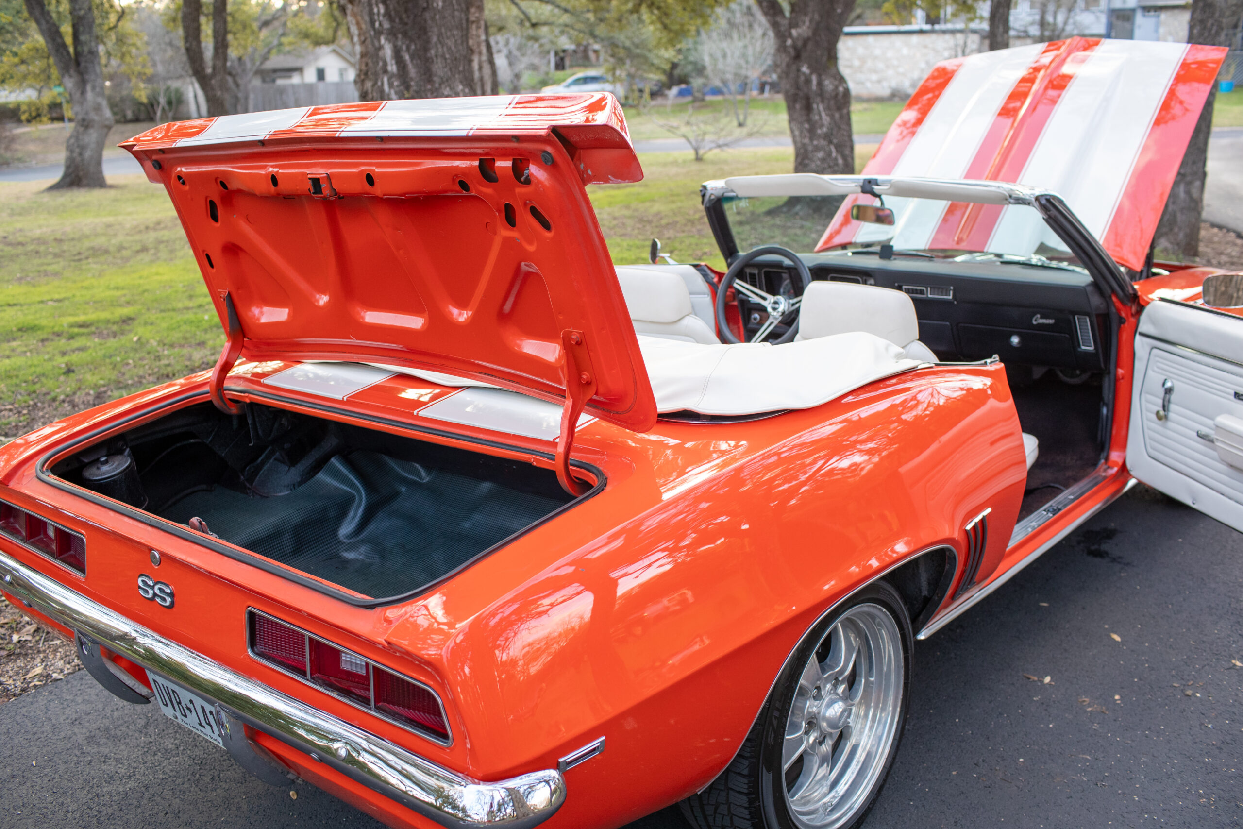 A classic orange convertible car with an open trunk and doors parked outdoors, featuring white racing stripes and a clean interior.