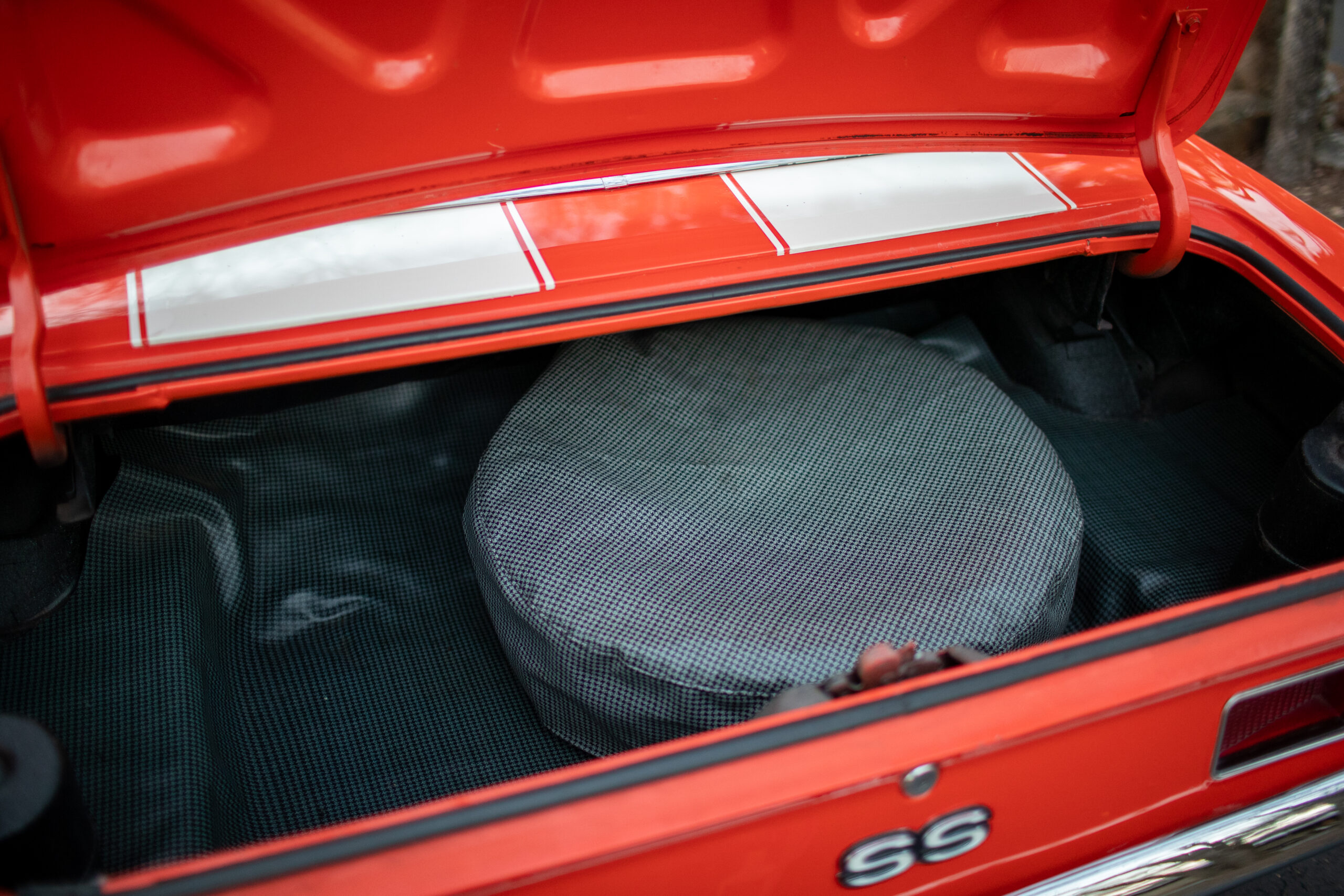 Open trunk of an orange car, revealing a spare tire covered with a gray, checkered fabric cover. The car's exterior has SS badges and white stripes.