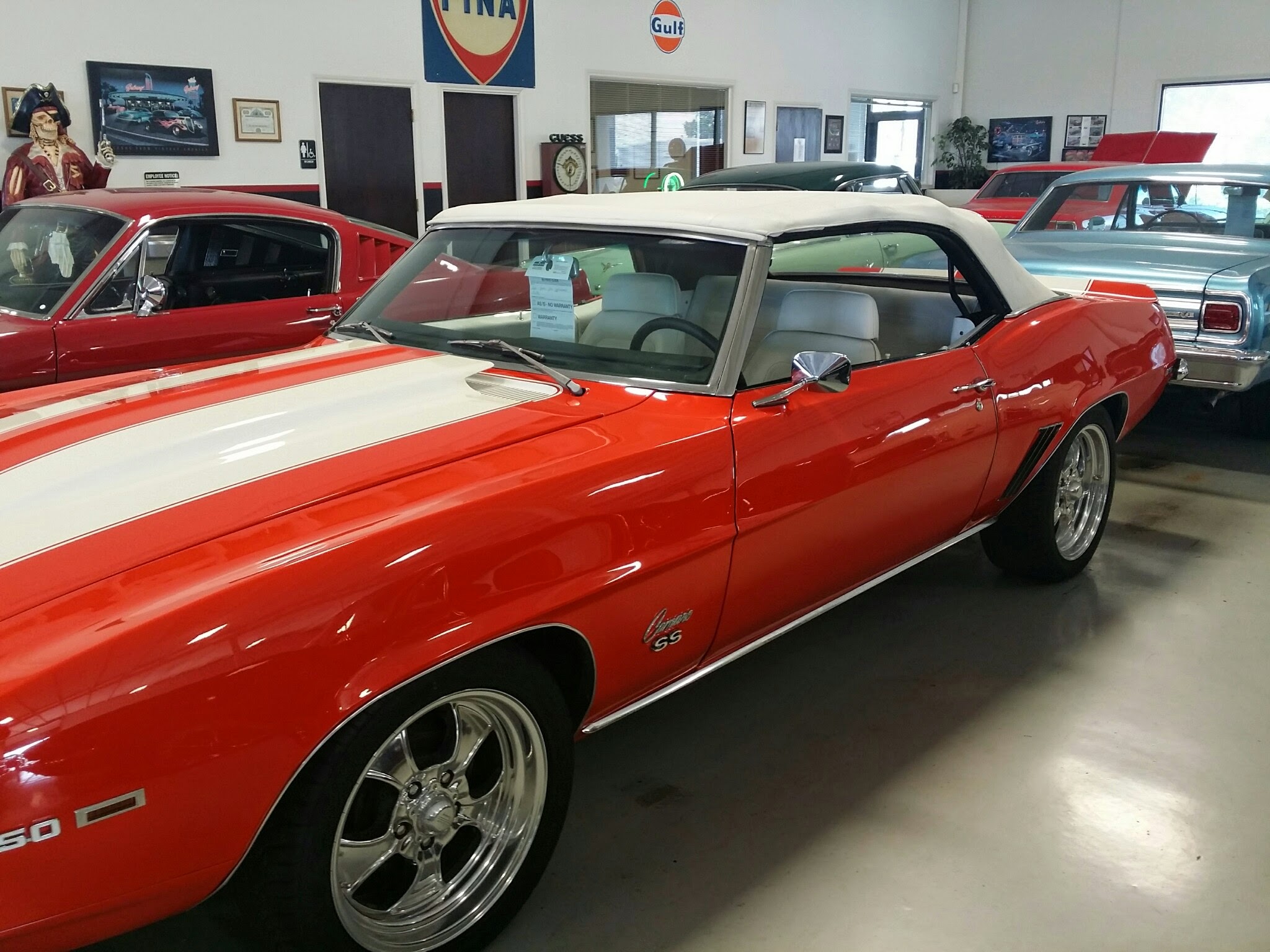 A classic red convertible with white racing stripes and polished chrome wheels parked indoors, surrounded by other vintage cars in a showroom.