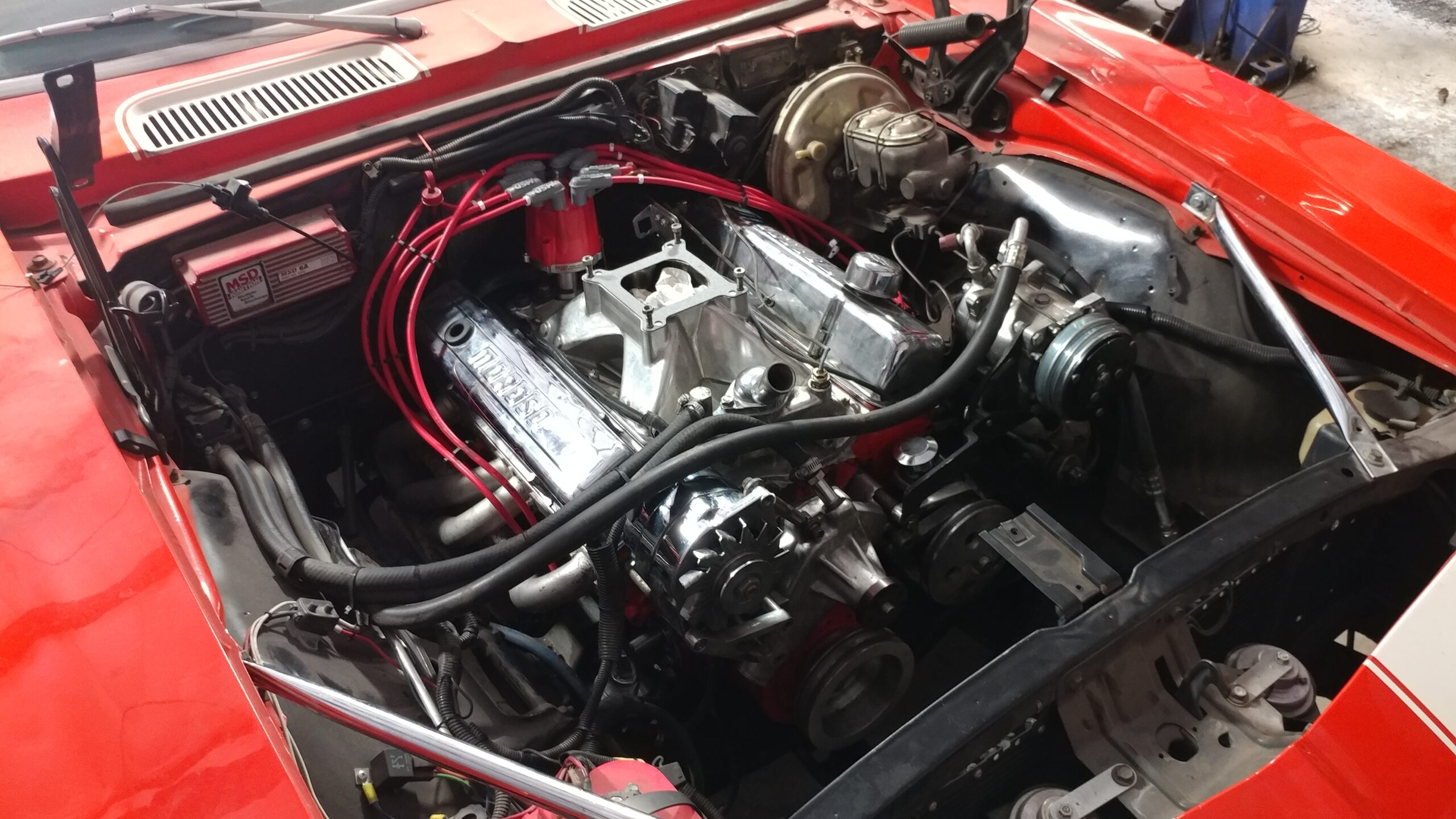 An open car hood reveals a V8 engine with various components and red ignition wires in a red vehicle.