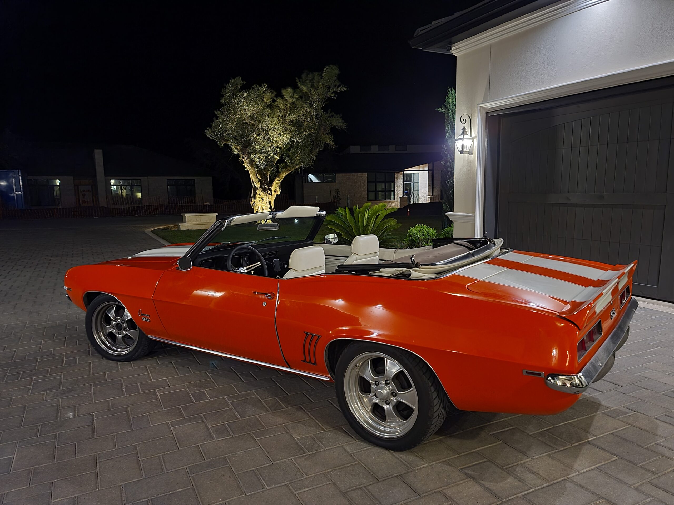 A red convertible car with white stripes is parked in a driveway at night. The house and garage are lit, and an illuminated tree is visible in the background.