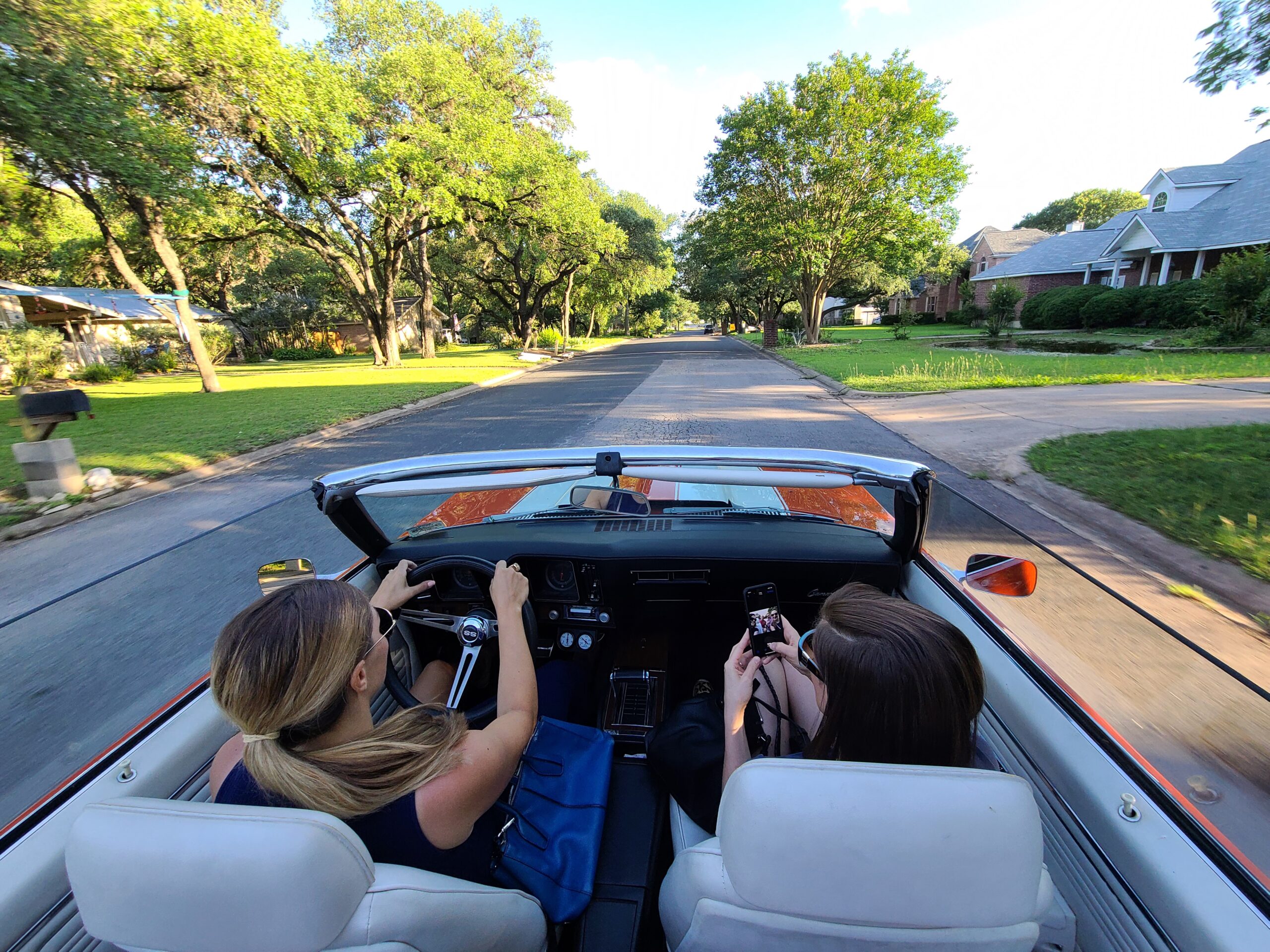 Two people are driving in a red convertible with the top down on a sunlit suburban street lined with trees and houses. The passenger is holding a camera and appears to be taking a photo.