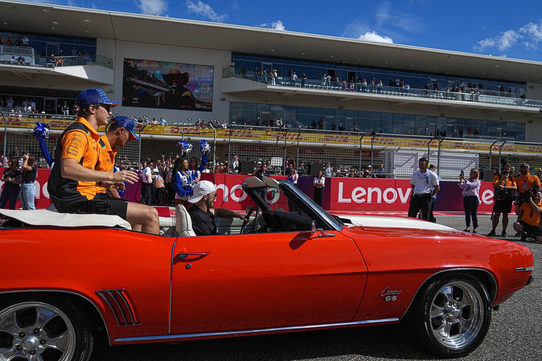 Two people sit in an orange convertible car at a racetrack event, with spectators and officials in the background. Two people wear matching orange and blue outfits.