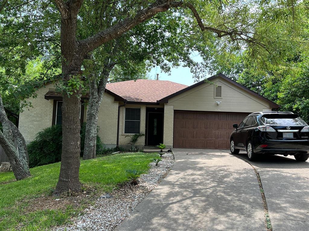 A single-story house with a beige exterior, a brown roof, and a wooden garage door, surrounded by trees and greenery. A black SUV is parked in the driveway.