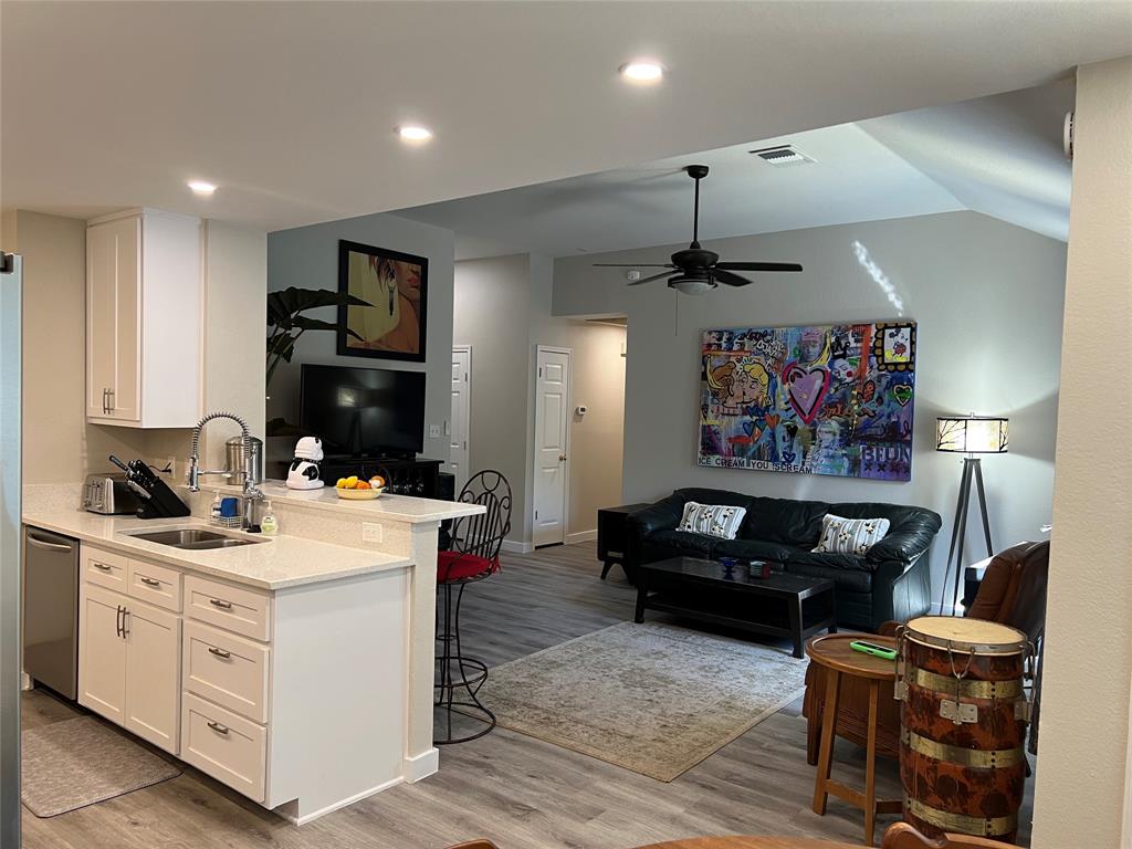 Open-concept living area with white kitchen cabinets, bar stools at the counter, black sofa, colorful wall art, flat-screen TV, ceiling fan, and a mix of wood and modern furnishings.