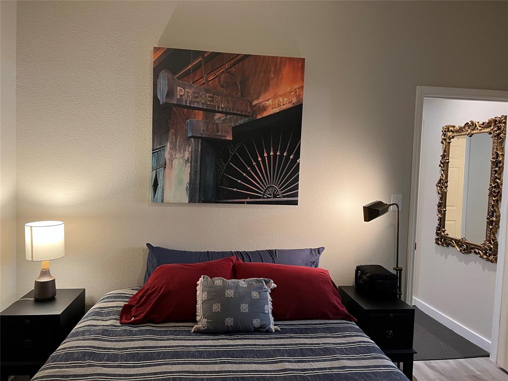 A bedroom with a striped bedspread, red and blue pillows, nightstands with lamps on either side, a large framed artwork above the bed, and a decorative mirror visible in the adjacent bathroom.