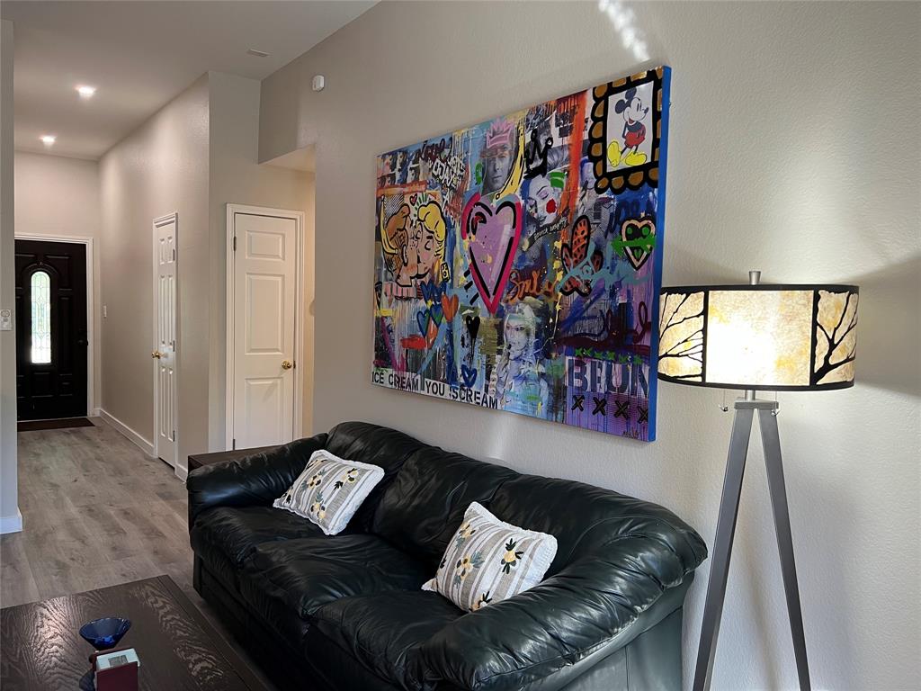 A living room with a black leather couch, two pillows, a colorful abstract painting on the wall, and a floor lamp beside the couch. A doorway is visible in the background.