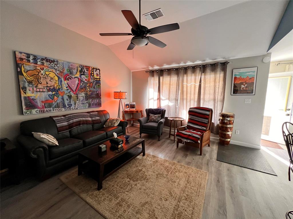 A cozy living room featuring a sofa, a coffee table, chairs, a large painting, a ceiling fan, and a window with curtains letting in natural light. The floor is covered with a light wood finish and a rug.