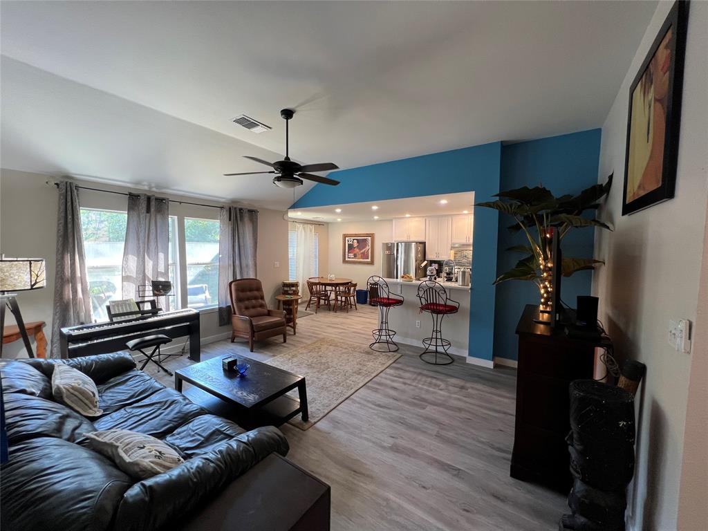 Modern living room with dark leather couch, light wood flooring, ceiling fan, and large windows. The room opens to a kitchen area with bar stools and dining table. Decor includes plants and artwork.