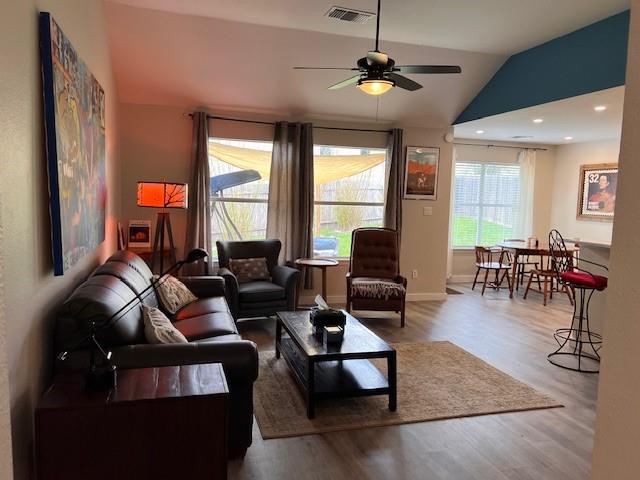 A living room with dark brown furniture, a rectangular coffee table, a ceiling fan, and large windows with grey curtains. The dining area has a round table and chairs near another large window.