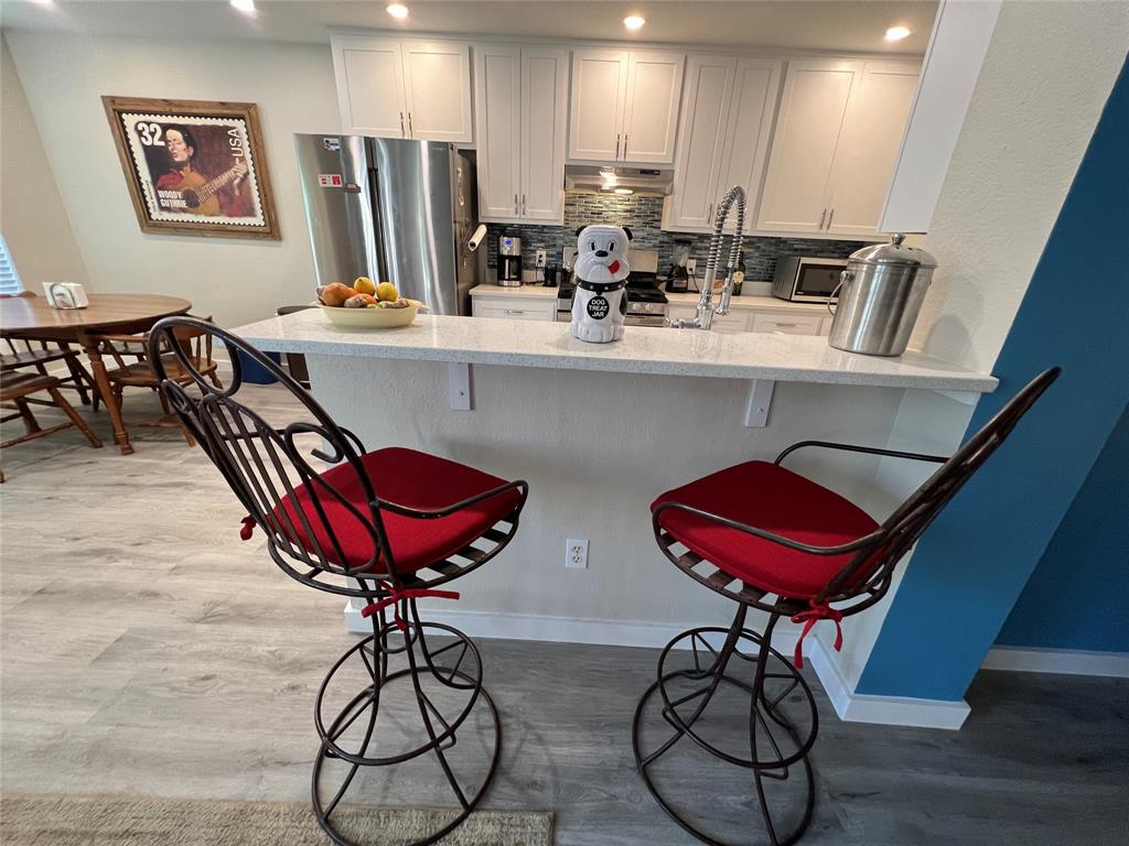 A kitchen with a countertop, two red cushioned bar stools, a fruit bowl, and a small dog-shaped coffee maker on the counter. The background features a stainless steel refrigerator and a dining area.