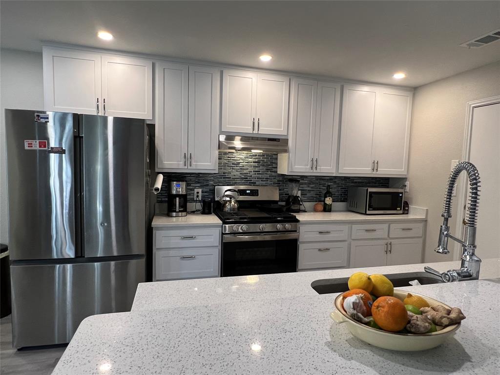 A modern kitchen with white cabinets, stainless steel appliances, and a fruit bowl on a quartz island countertop. The backsplash features a tiled design, and a high-arc faucet is present.