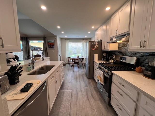 A modern kitchen with white cabinetry, stainless steel appliances, and grey flooring leads to a dining area with a wooden table and chairs by large windows.